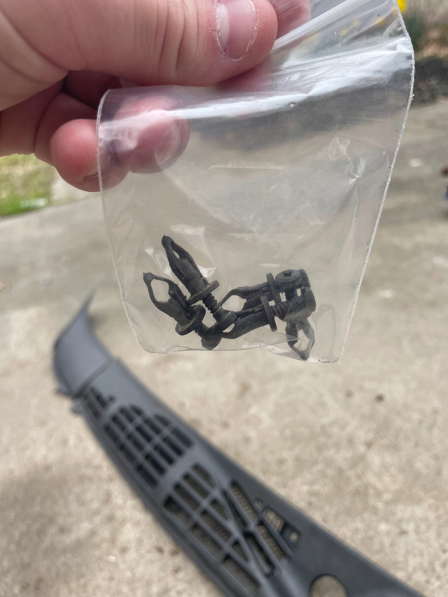A hand holds a clear plastic bag of black fasteners for the OEM Wiper Cowl Cover Set (1994-2005 Chevrolet S10, Blazer, and more) by General Motors, with a Chevrolet Blazer grille and concrete ground in the background.