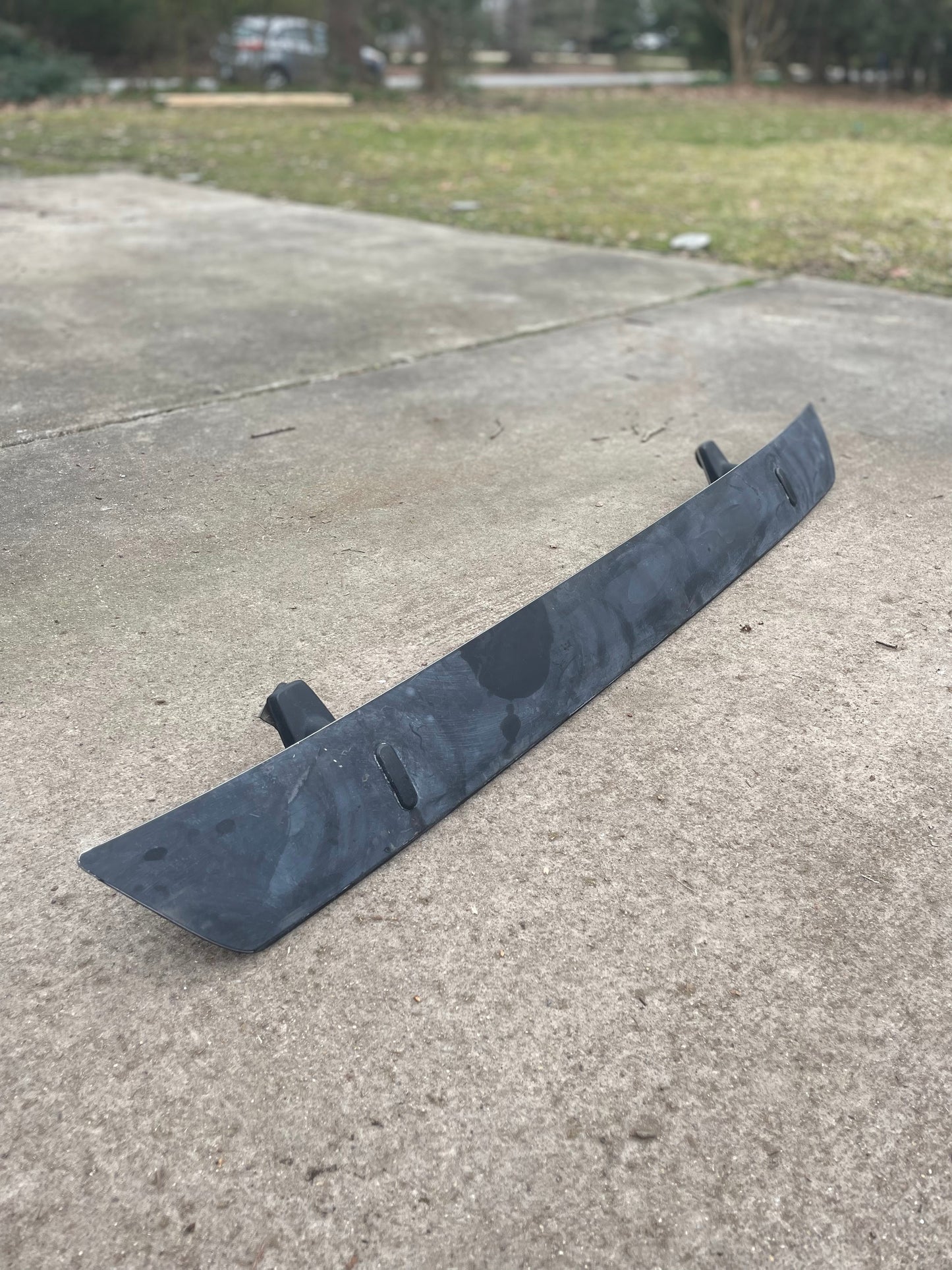 A pair of black OEM Rear Hatch Hinges with Spoiler Wing for 1983-1994 Chevrolet S10 Blazer by General Motors lies on a concrete driveway, with grass and trees blurred in the background.