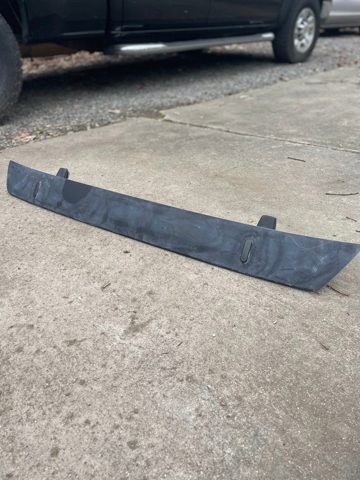 An OEM Rear Hatch Hinges with Spoiler Wing by General Motors for 1983-1994 Chevrolet S10 Blazer lies on a concrete driveway, with a black vehicle and gravel seen in the background.