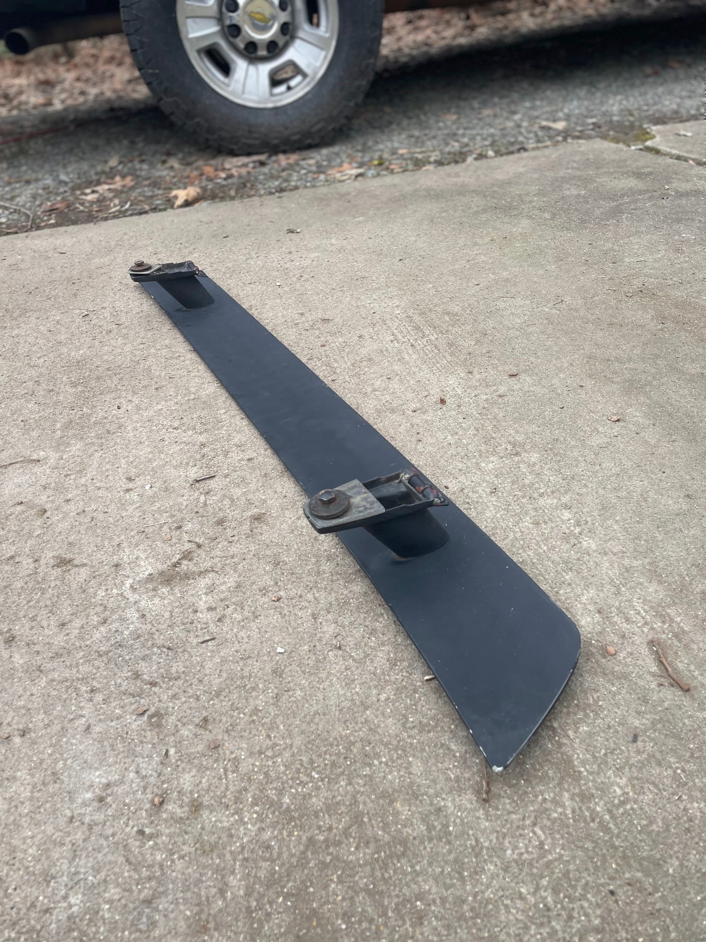 A single metal lawn mower blade with two bolts rests on concrete. In the blurred background, a tire and a vehicle are visible, possibly featuring General Motors OEM Rear Hatch Hinges with Spoiler Wing for 1983-1994 Chevy S10 Blazer.