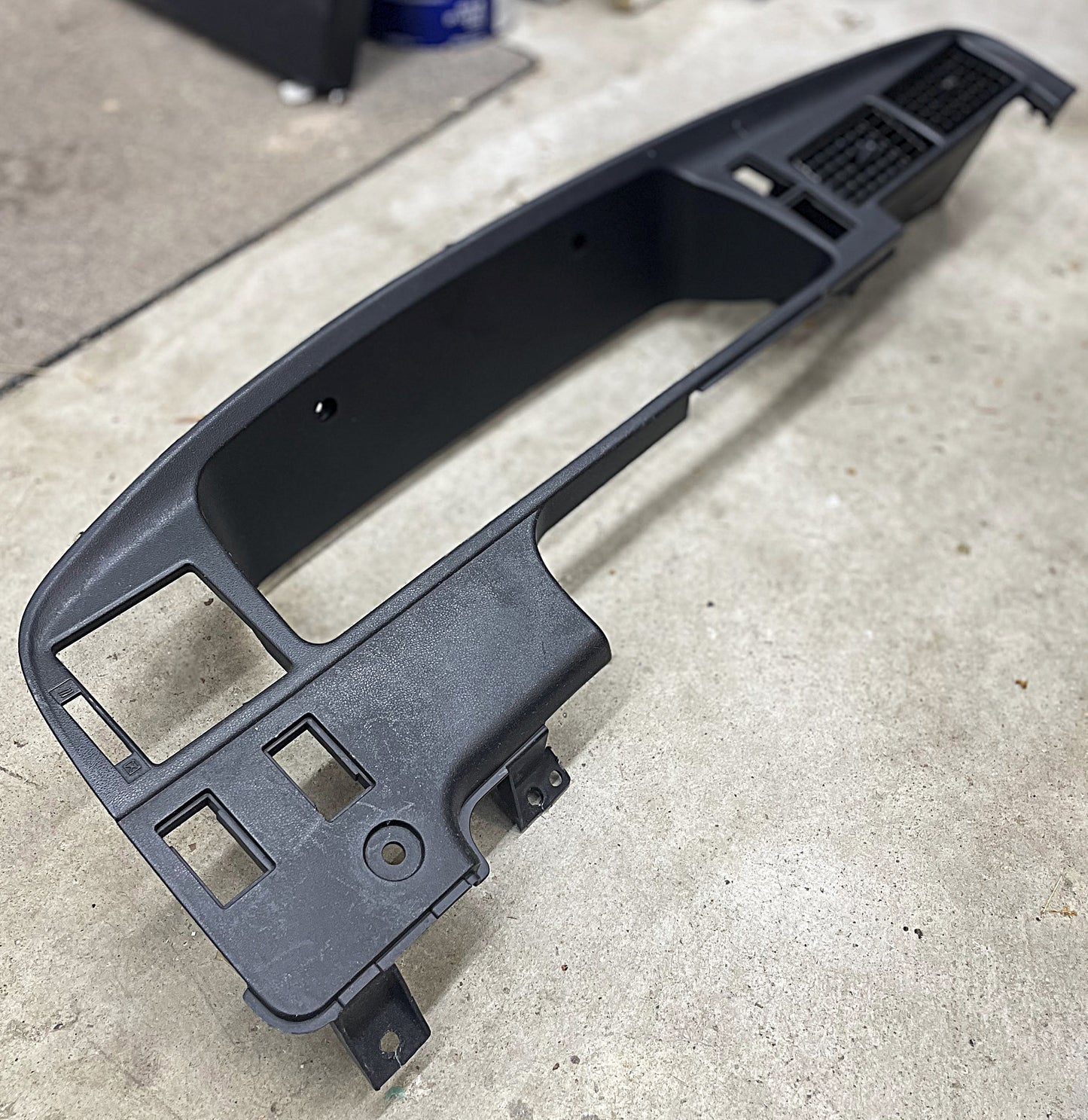 A General Motors OEM Instrument Cluster Dash Bezel for 1989-1995 Toyota Pickup and 4Runner rests on a concrete floor, featuring rectangular gauge, vent, and control cutouts. Authentic part removed from a vehicle for repair or replacement.