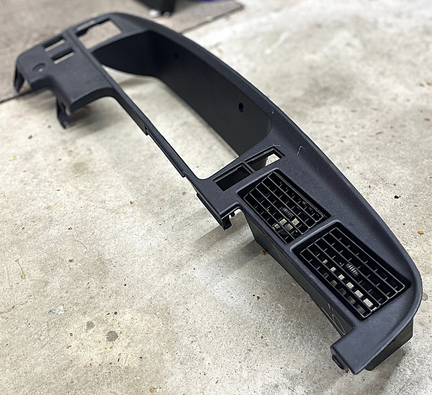 An OEM Instrument Cluster Dash Bezel for 1989-1995 Toyota Pickup or 4Runner by General Motors is shown on a concrete floor, detached and with air vents visible, possibly ready for repair or installation.
