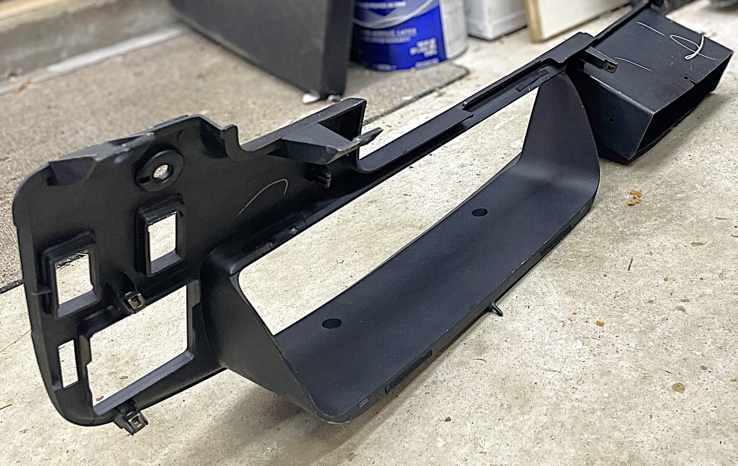 A General Motors OEM Instrument Cluster Dash Bezel for 1989-1995 Toyota Pickup & 4Runner, black plastic with cutouts and compartments, rests on a concrete garage floor with storage items in the background.
