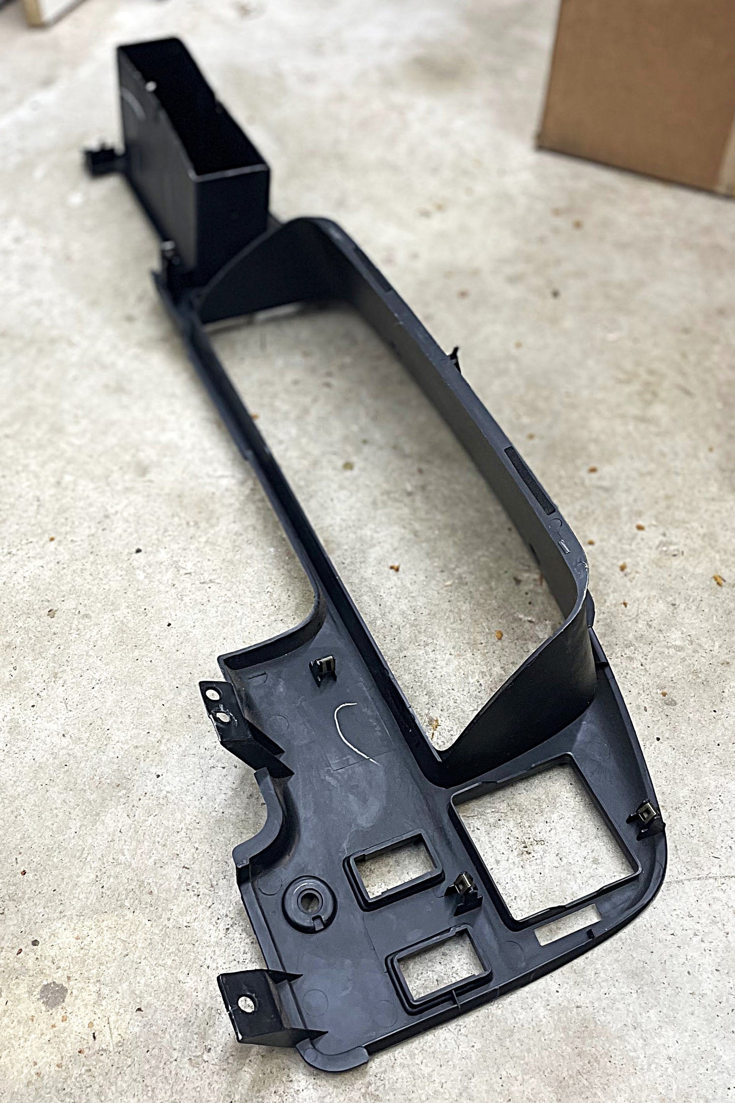 A General Motors OEM Instrument Cluster Dash Bezel for 1989-1995 Toyota Pickup and 4Runner in black plastic is laying on a light gray concrete floor, with a cardboard box partly visible in the background.