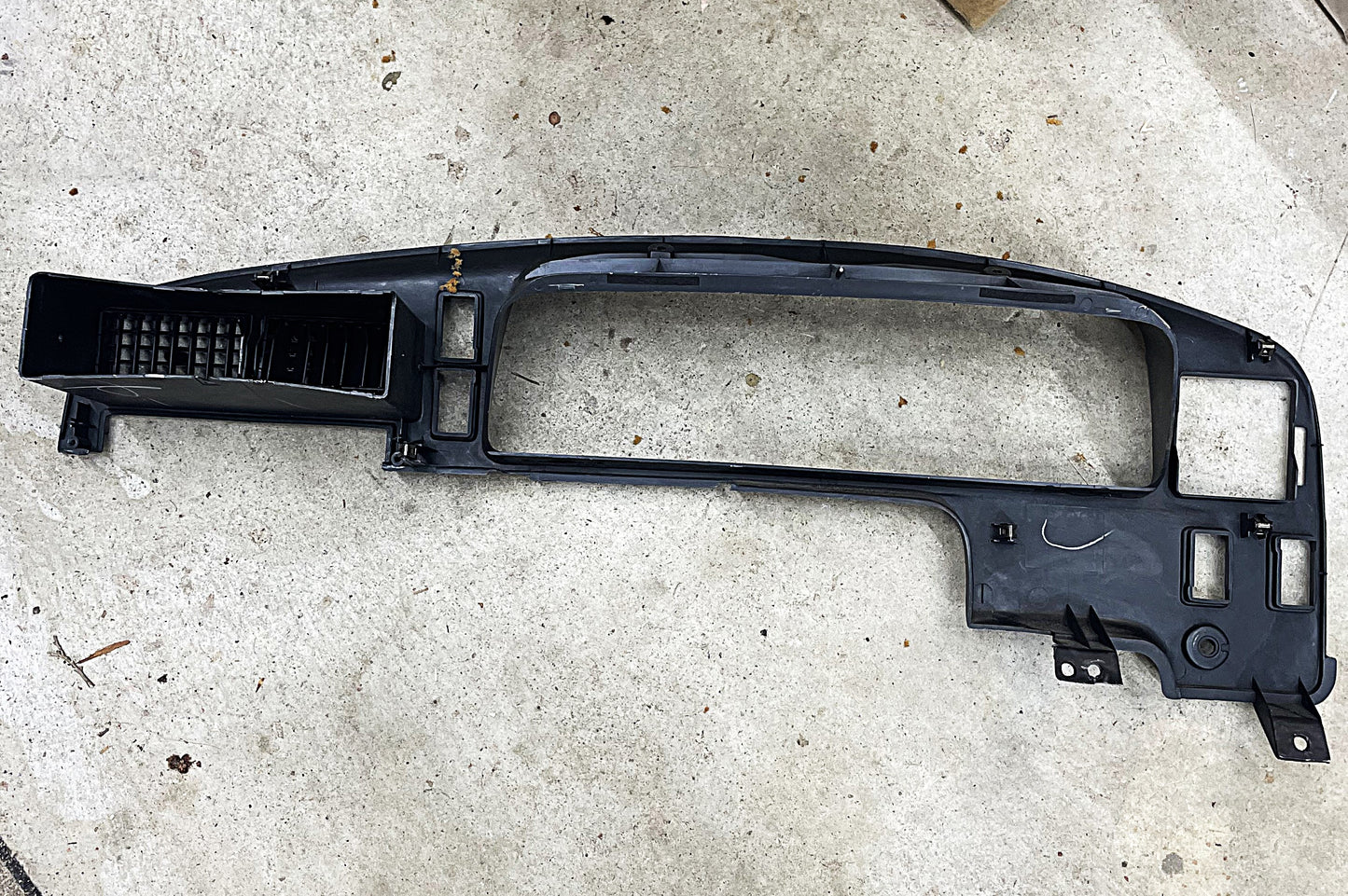 An OEM Instrument Cluster Dash Bezel for 1989-1995 Toyota Pickup, 4Runner by General Motors rests on concrete, featuring vent, gauge, and control cutouts with minor wear and dust from previous interior use.