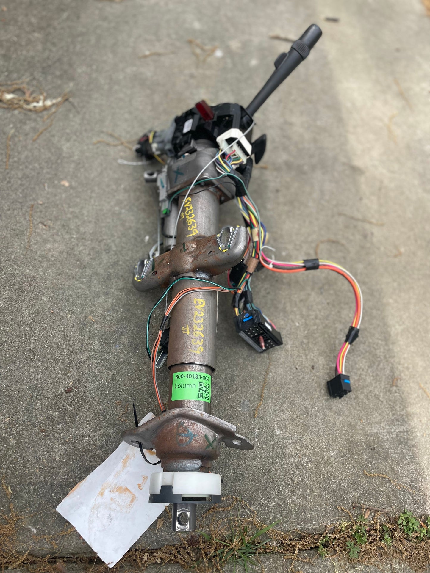 A General Motors OEM Steering Column w/ KEY for 2000-2002 GM Full Size Trucks, featuring wiring harnesses and connectors for automatic transmission, is shown disassembled on a concrete surface with visible colored wires and labels.