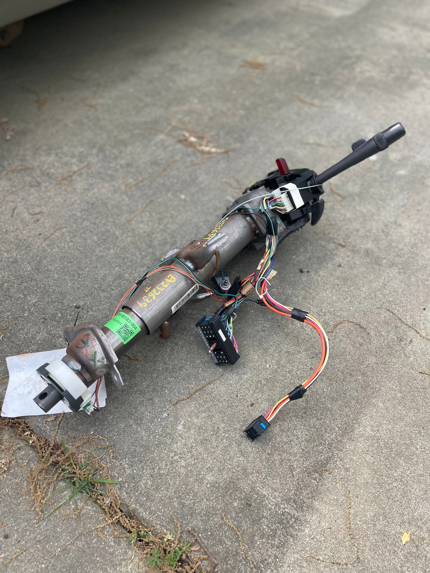 A General Motors OEM Steering Column w/KEY for 2000-2002 GM Full Size Trucks, with attached wiring harnesses and connectors, is shown outdoors on concrete. Various labels and wires are visible on the automatic transmission component.