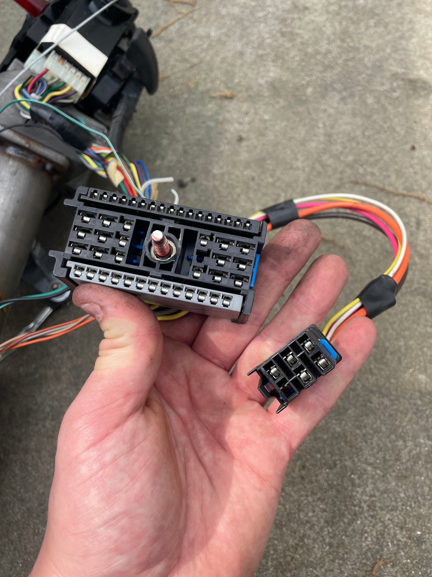 A hand holds two General Motors automotive connectors with colored wires on concrete. The larger fits an OEM Steering Column w/KEY for 2000-2002 GM Full Size Trucks; the smaller has metal pins, likely for a Chevrolet Silverado transmission.