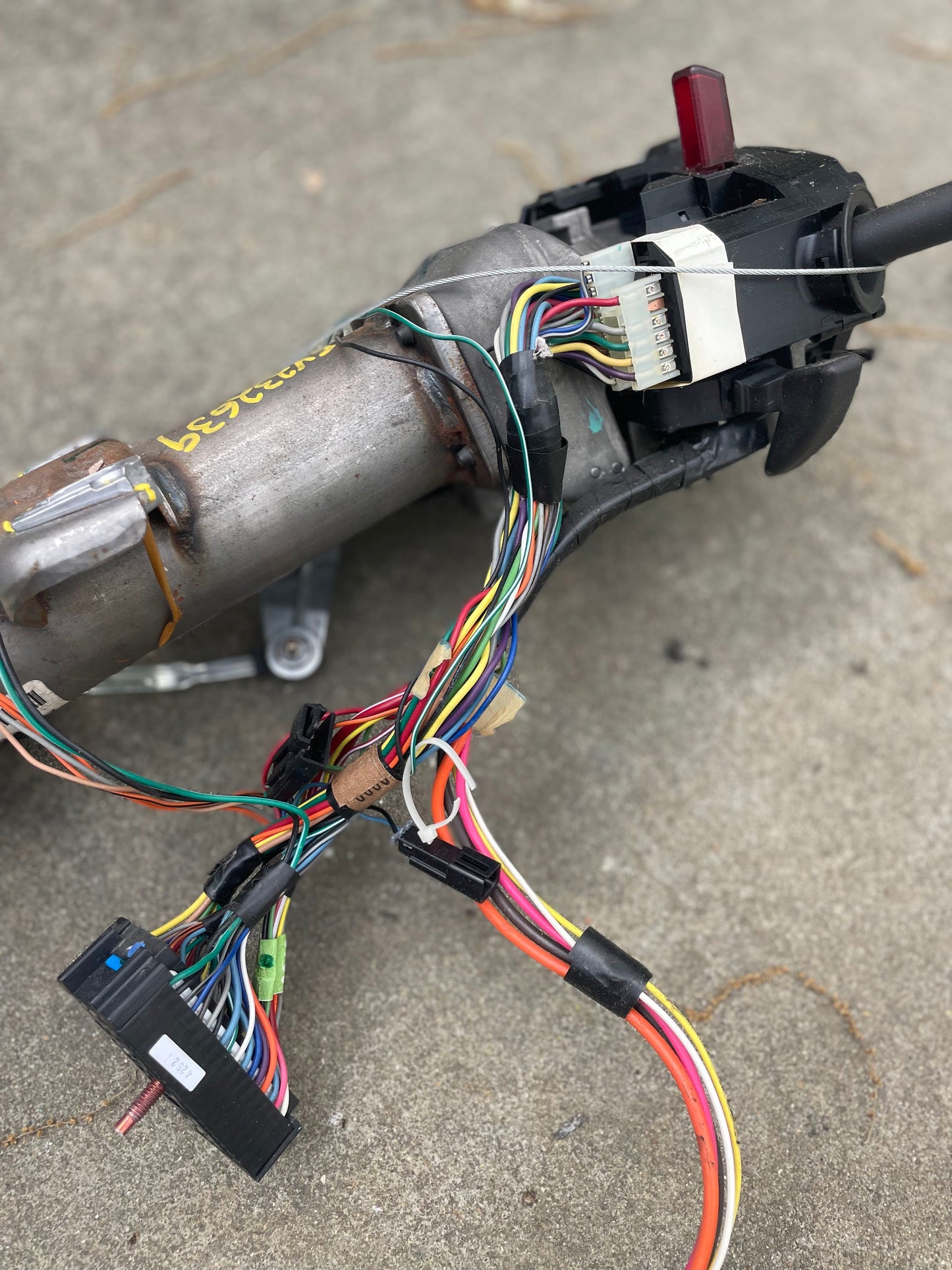 A close-up shows a General Motors OEM Steering Column w/KEY for 2000-2002 GM Full Size Trucks with colorful wires and connectors exposed on concrete. Some components are labeled, and the background is blurred.