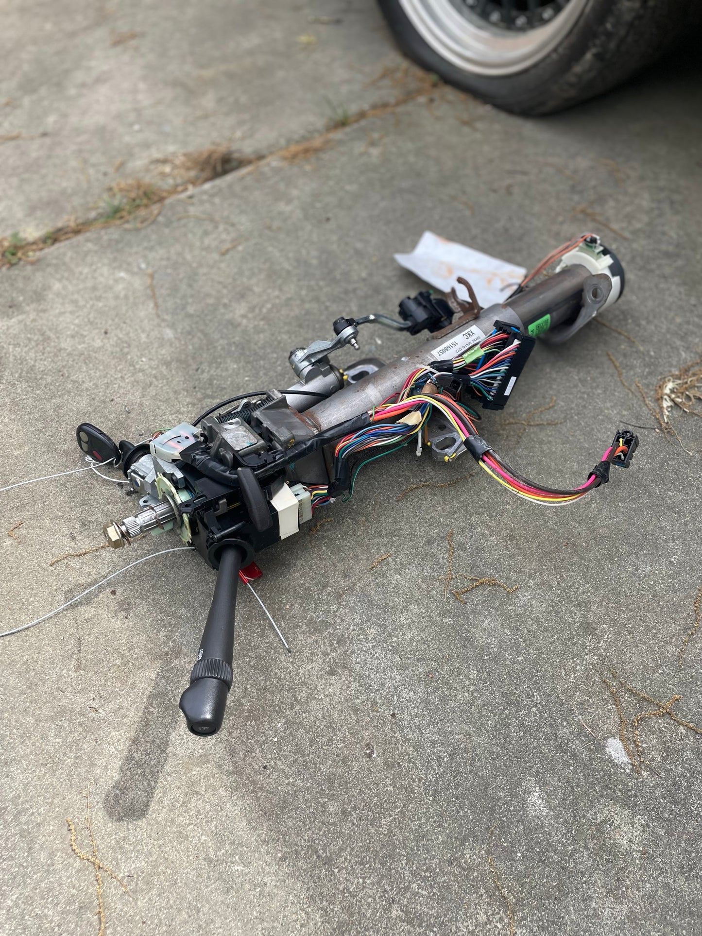 A General Motors OEM Steering Column w/KEY for 2000-2002 GM Full Size Trucks, with exposed wiring and levers, is shown disassembled on a concrete surface near a vehicle tire.