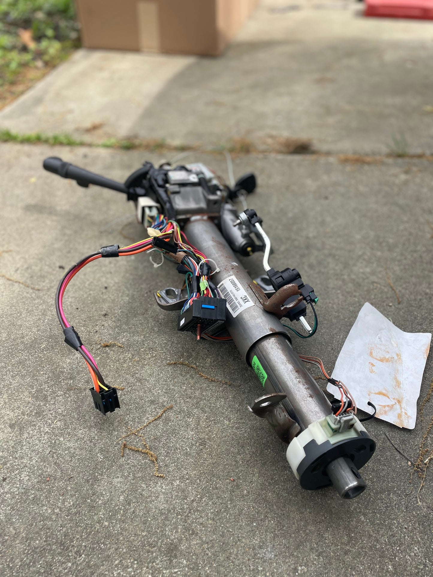 A General Motors OEM Steering Column w/KEY for 2000-2002 GM Full Size Trucks, featuring wiring and electronic parts, is on a concrete surface outdoors, with a white paper and several cardboard boxes in the background.