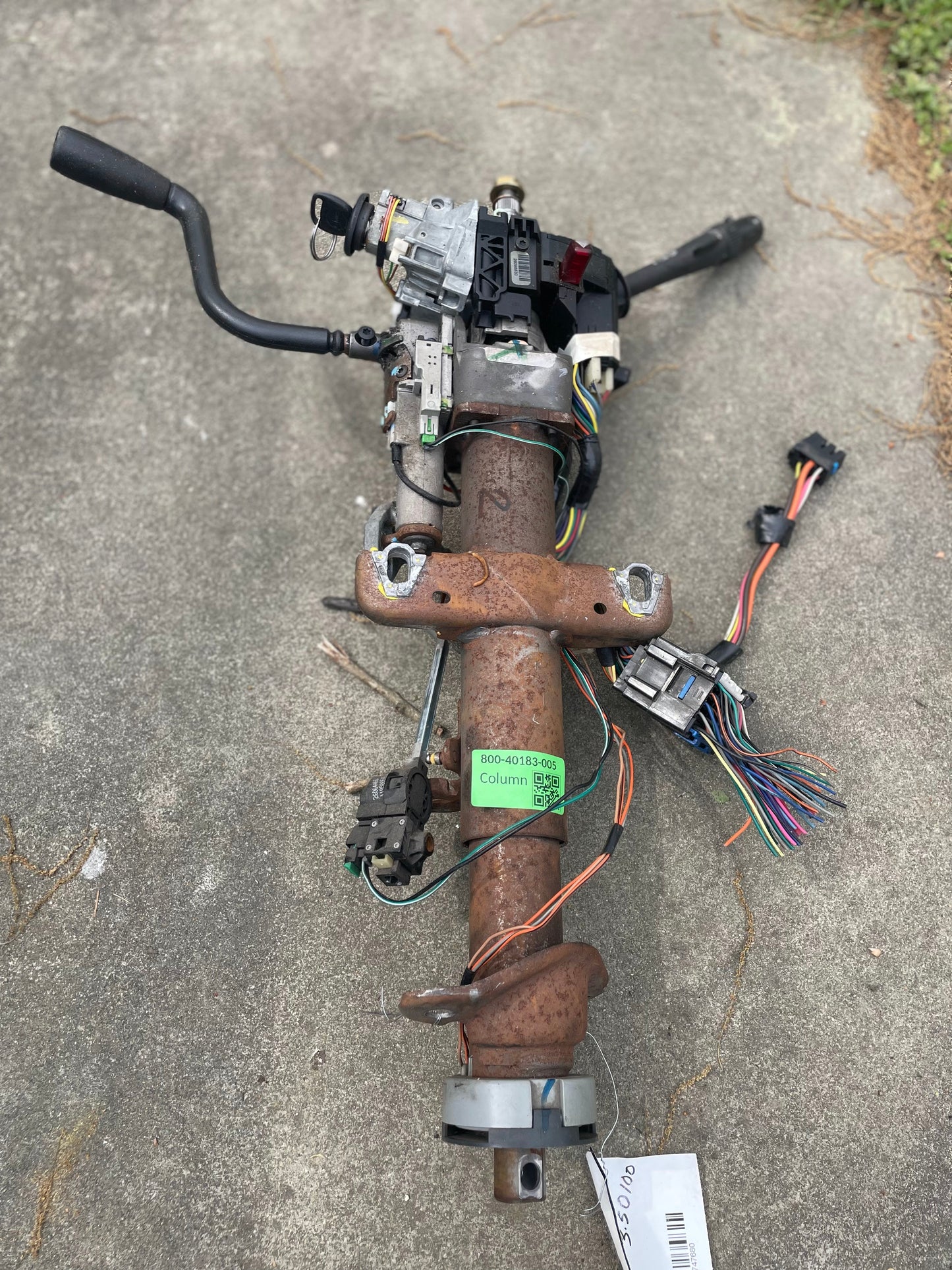 A rusty General Motors OEM Steering Column w/KEY for 2000-2002 GM Full Size Trucks, featuring attached wiring, sensors, connectors, and various colored wires for automatic transmission, rests on a concrete surface.