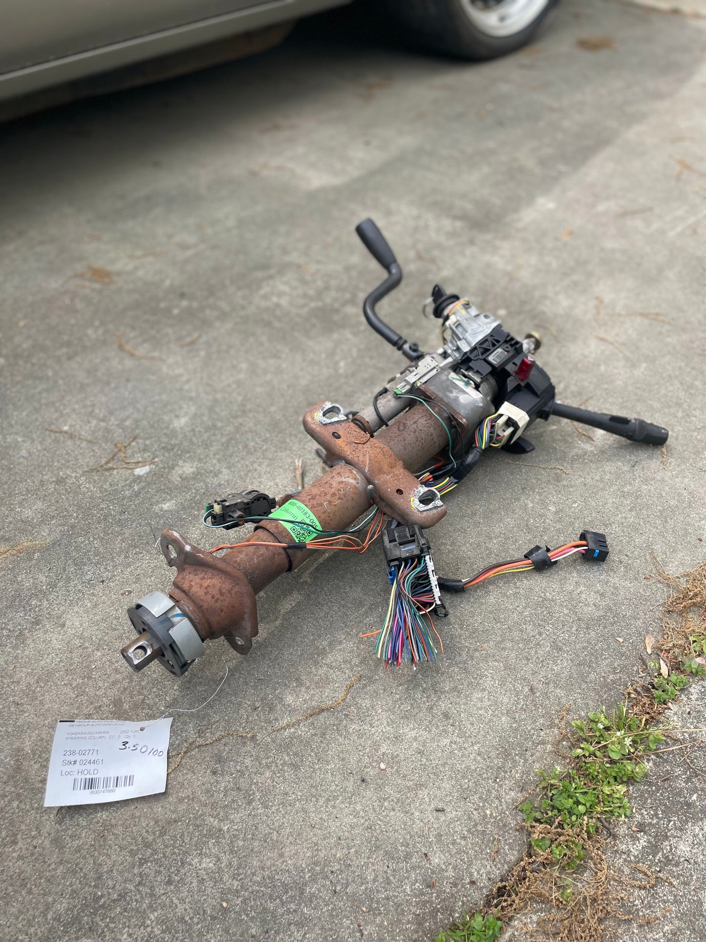 A General Motors OEM Steering Column w/KEY for 2000-2002 GM Full Size Trucks rests on a concrete driveway with wiring and components attached. A handwritten note is nearby, and a car tire is partly visible in the background.