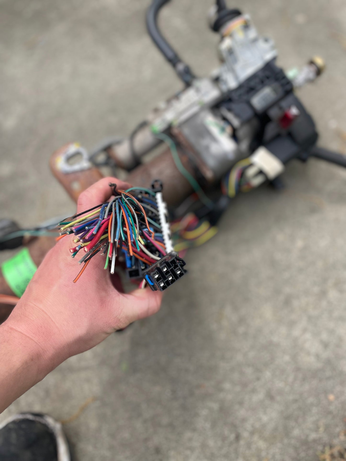 A hand holds the General Motors OEM Steering Column w/KEY for 2000-2002 GM Full Size Trucks, with colorful wires and a black connector visible; an automatic transmission part appears in the background on concrete.
