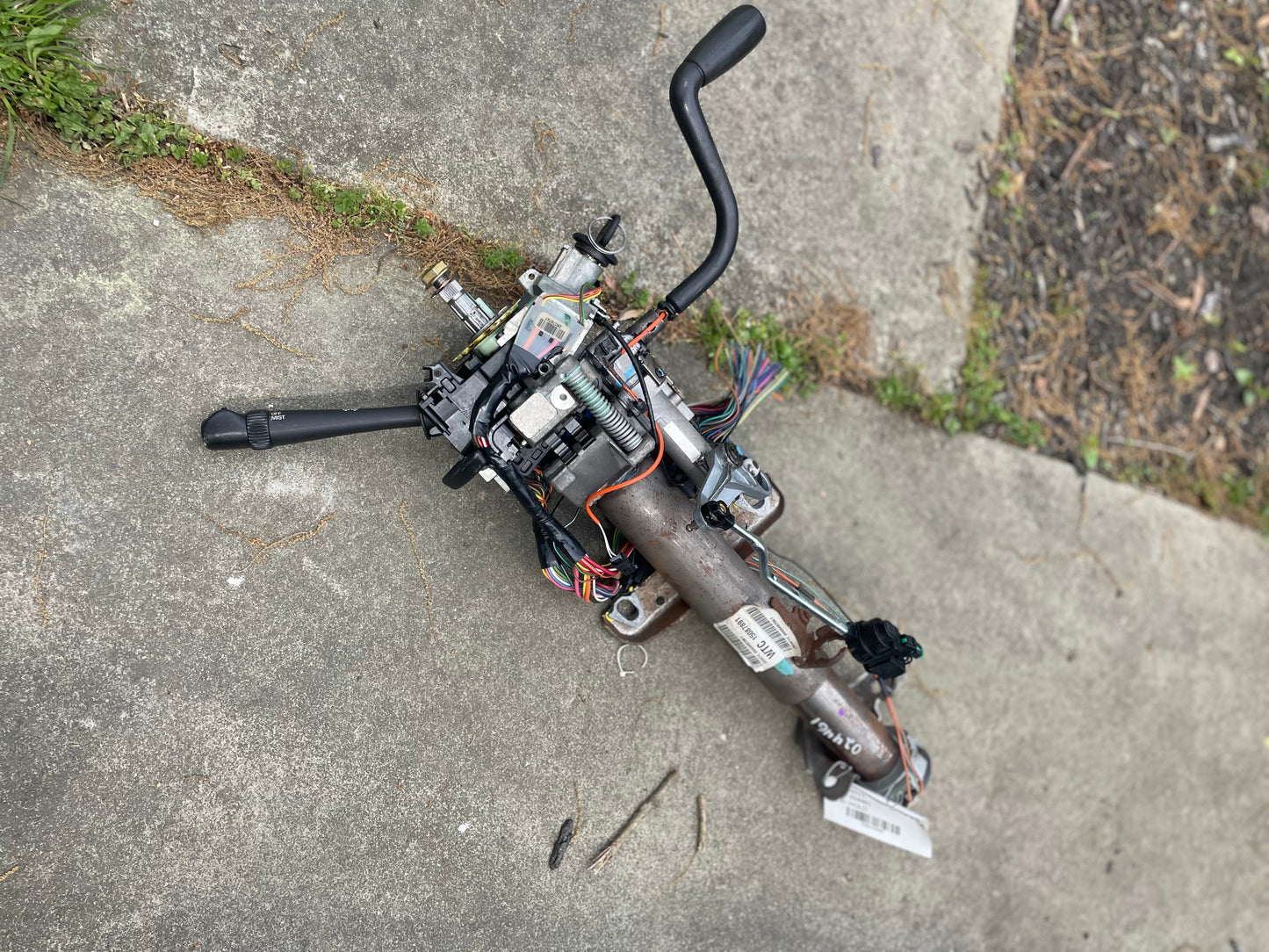 A General Motors OEM Steering Column w/KEY for 2000-2002 GM Full Size Trucks, with wiring and levers, rests outdoors on concrete near scattered screws and a patch of grass and soil.