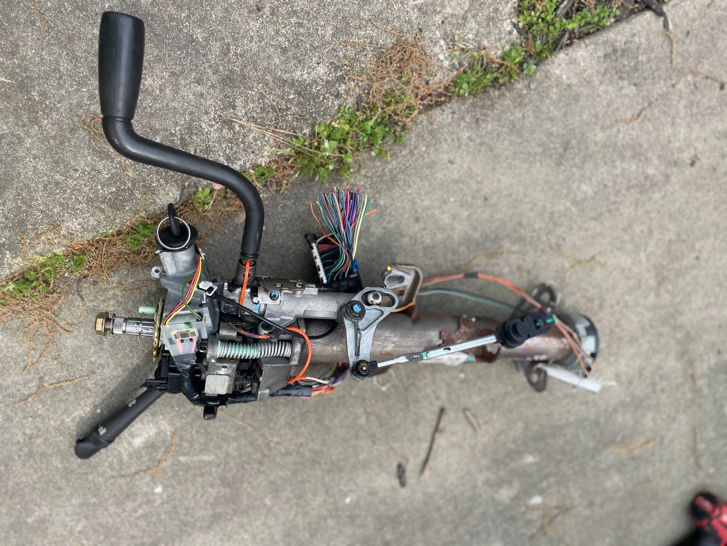 A General Motors OEM Steering Column w/KEY for 2000-2002 GM Full Size Trucks lies disassembled on concrete amid grass and debris, showing exposed wires and mechanical parts.