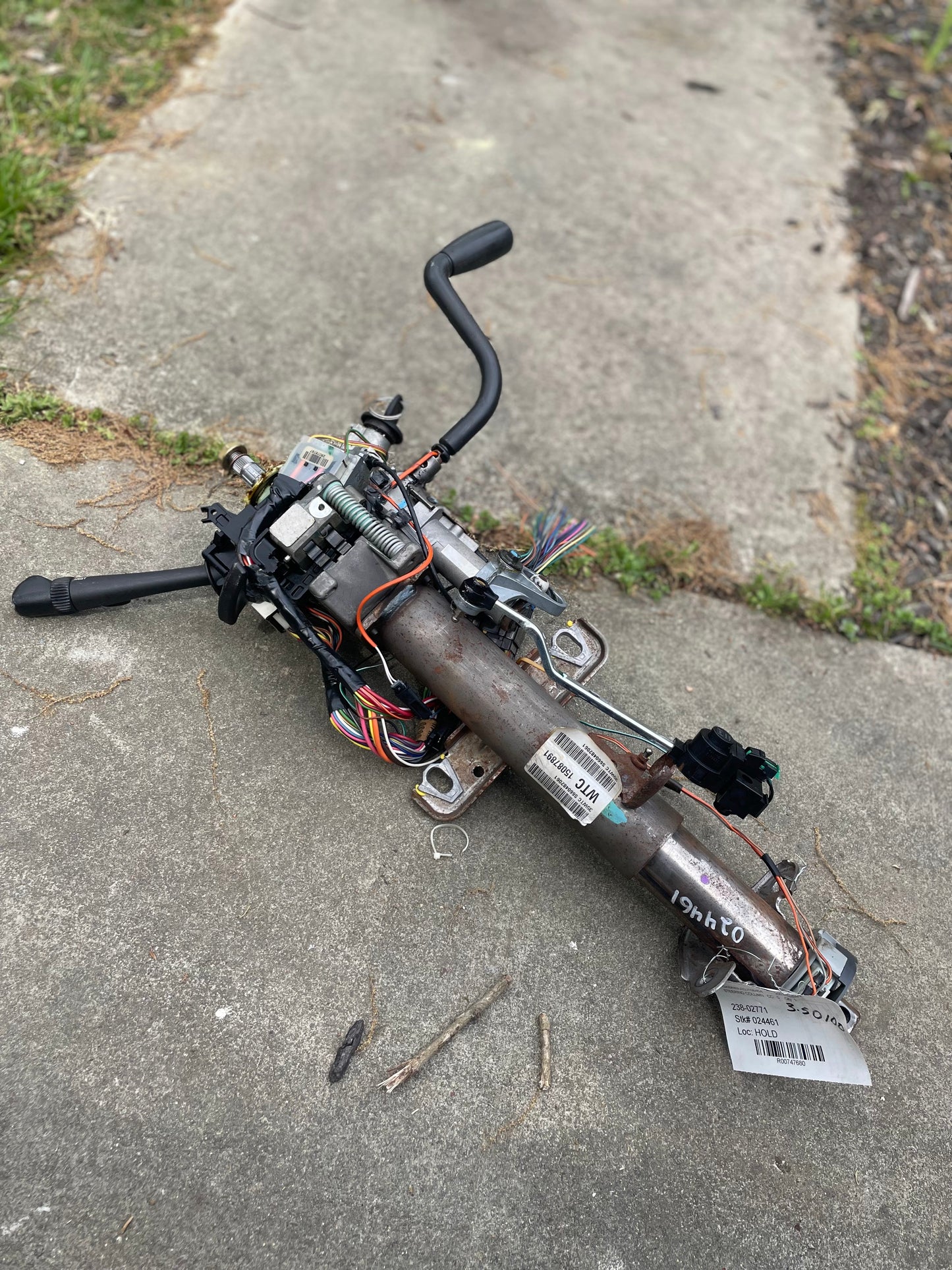 A General Motors OEM Steering Column w/KEY for 2000-2002 GM Full Size Trucks, with wiring and control levers attached, is disassembled and lying on a concrete sidewalk beside grass.