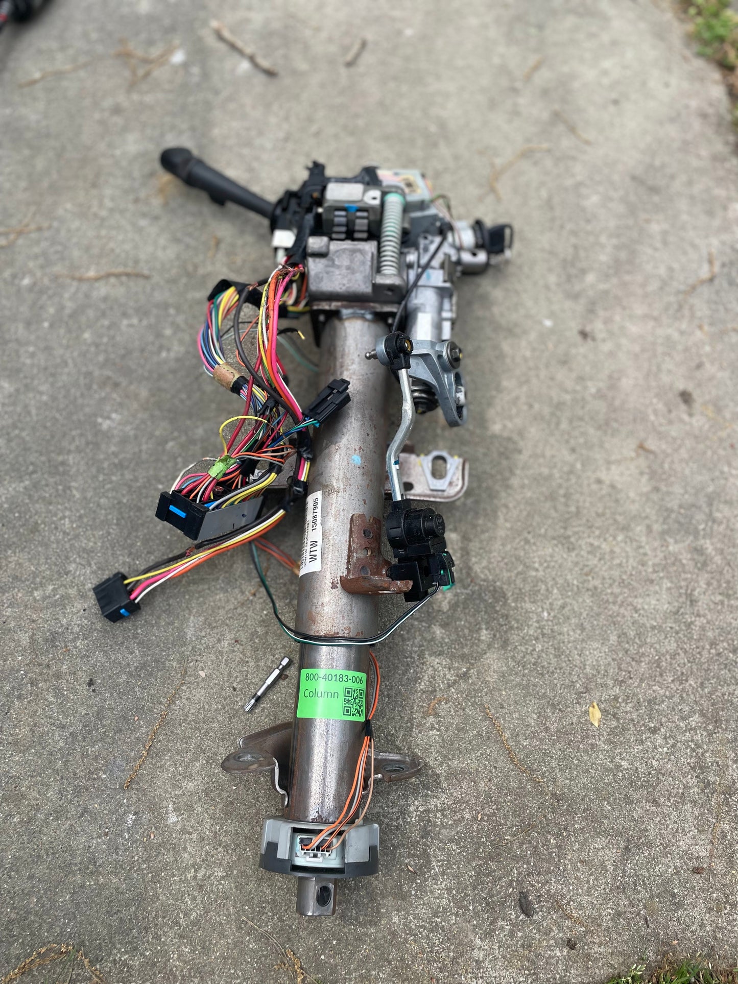 A General Motors OEM Steering Column w/KEY for 2000-2002 GM Full Size Trucks is shown outdoors on a concrete surface, with wiring harness, connectors, mechanical parts attached, a green label, and multicolored wires visible on the metal column.
