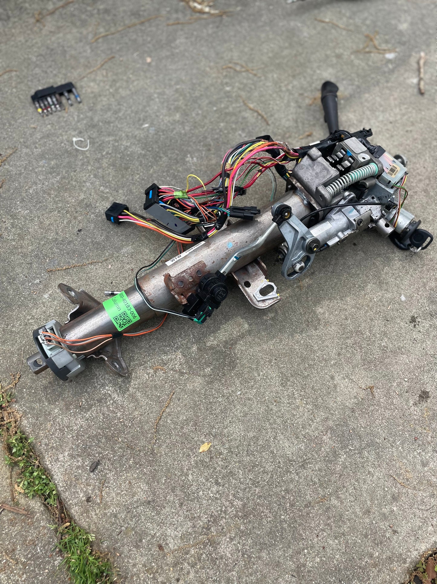 A General Motors OEM Steering Column w/KEY for 2000-2002 GM Full Size Trucks, with wiring and connectors attached, is shown on a concrete surface outdoors; small tools are visible in the background.