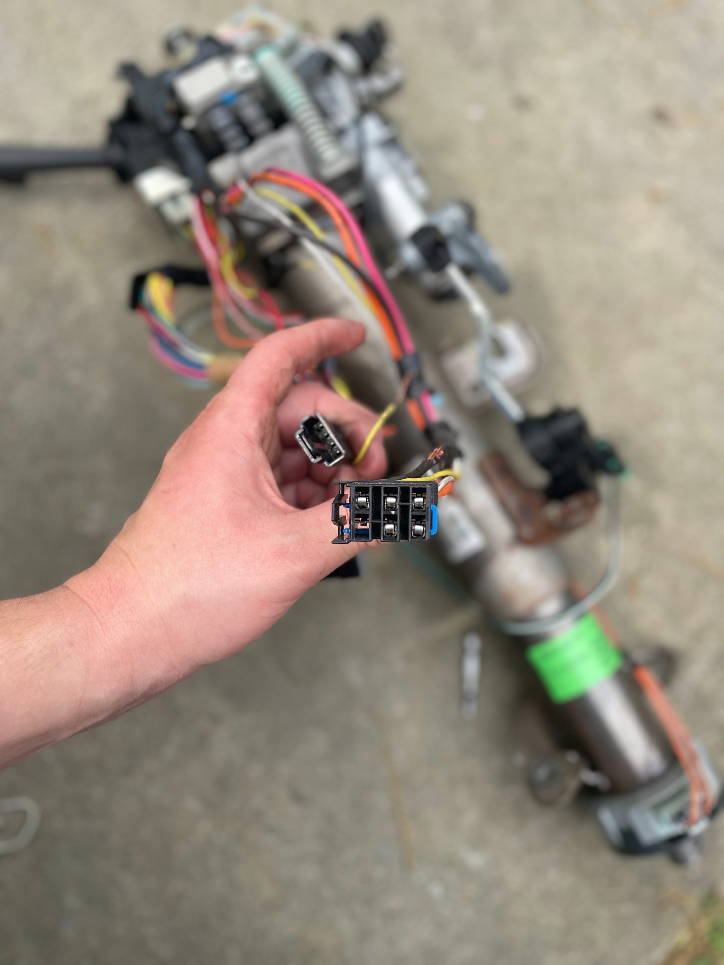 A hand holds two electrical connectors above a General Motors OEM Steering Column w/KEY for 2000-2002 GM Full Size Trucks, showing exposed colored wires and components on a concrete surface.