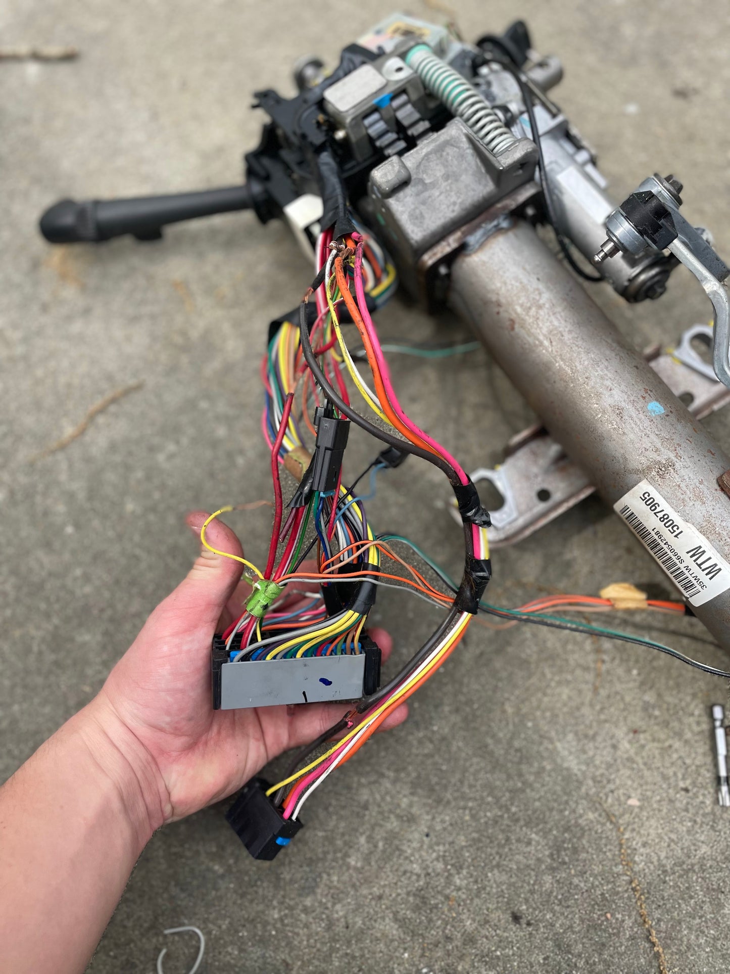 A hand holds colorful electrical wires attached to a General Motors OEM Steering Column w/KEY for 2000-2002 GM Full Size Trucks, resting on a concrete surface.