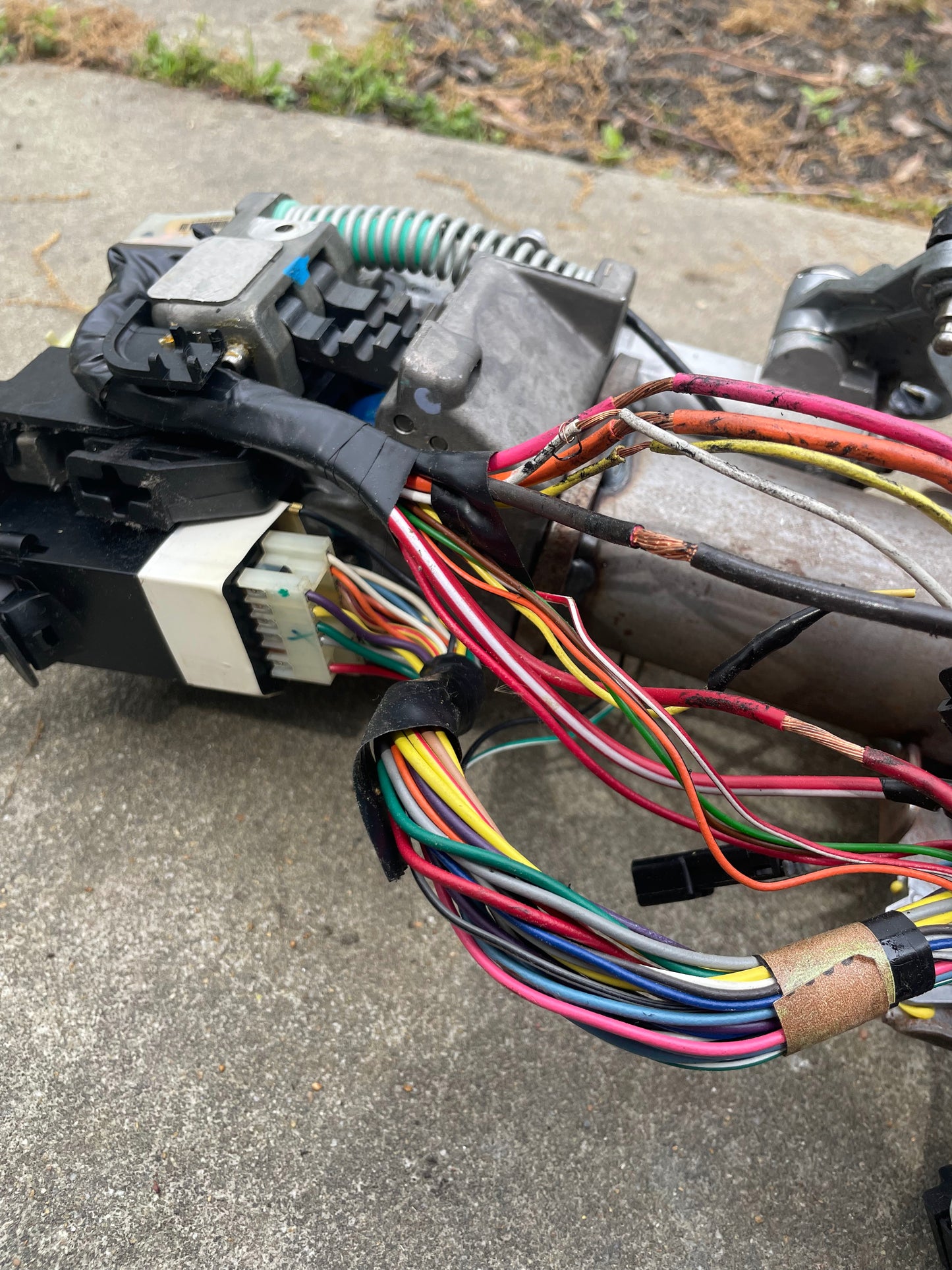 A close-up of colorful electrical wires and connectors attached to a metal component from a General Motors OEM Steering Column w/KEY for 2000-2002 GM Full Size Trucks, resting on a concrete surface outdoors.