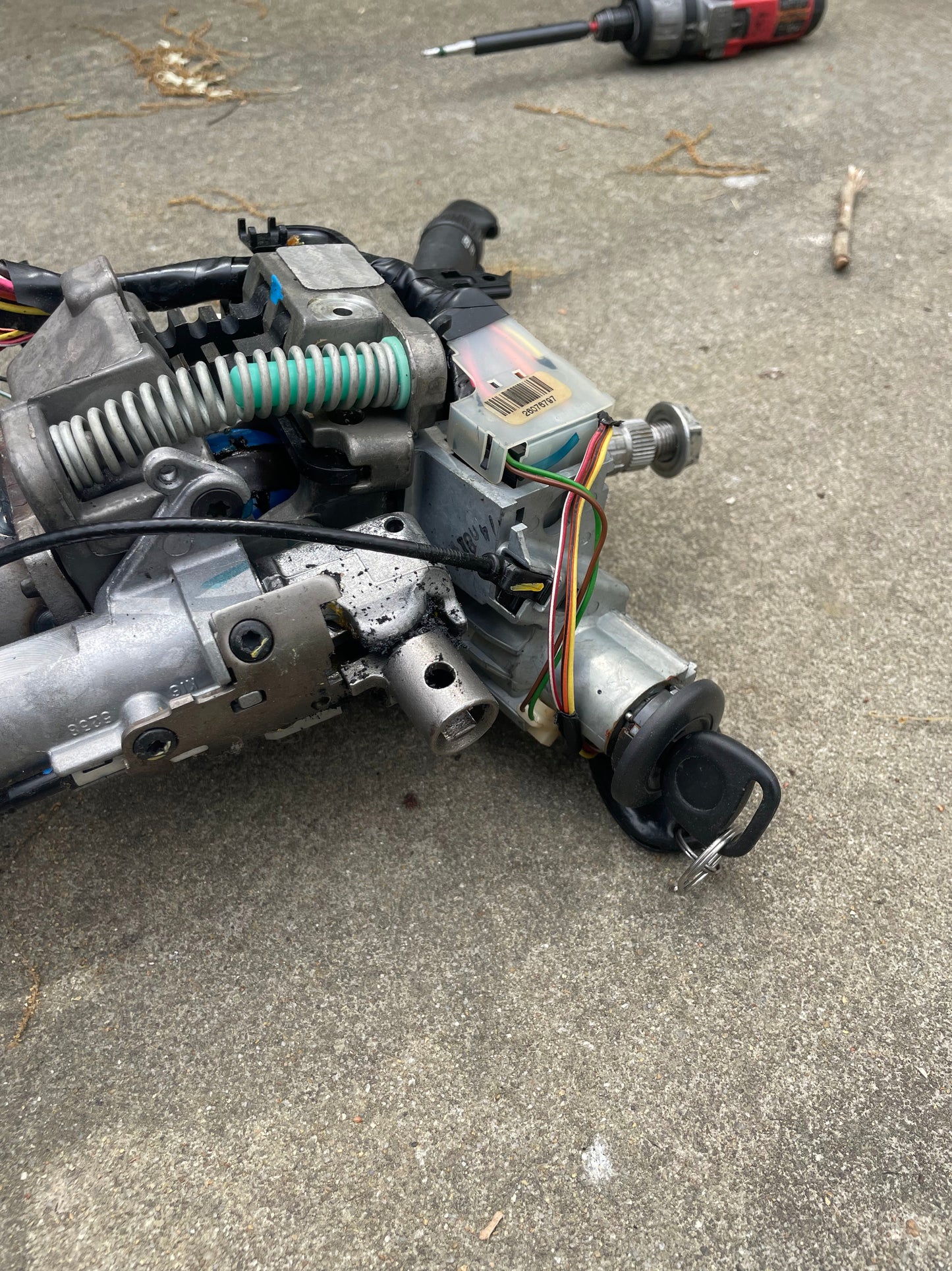 A close-up of a disassembled General Motors OEM Steering Column w/KEY for 2000-2002 GM Full Size Trucks shows exposed wires and the key in the ignition switch, resting on concrete with a screwdriver and stick in the background.