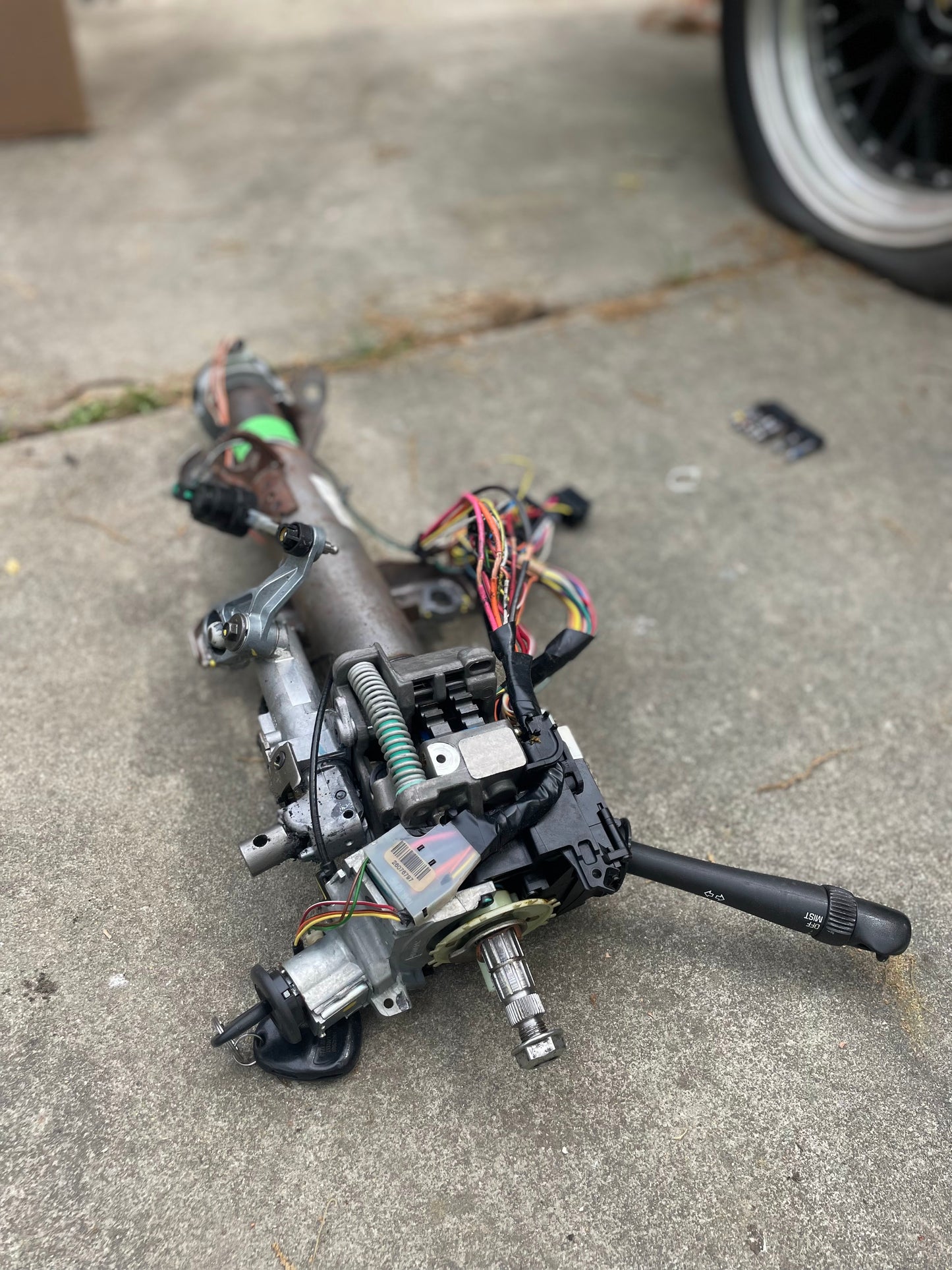 A General Motors OEM Steering Column w/KEY for 2000-2002 GM Full Size Trucks, made for automatic transmission, is outdoors on concrete beside a tire and grass.