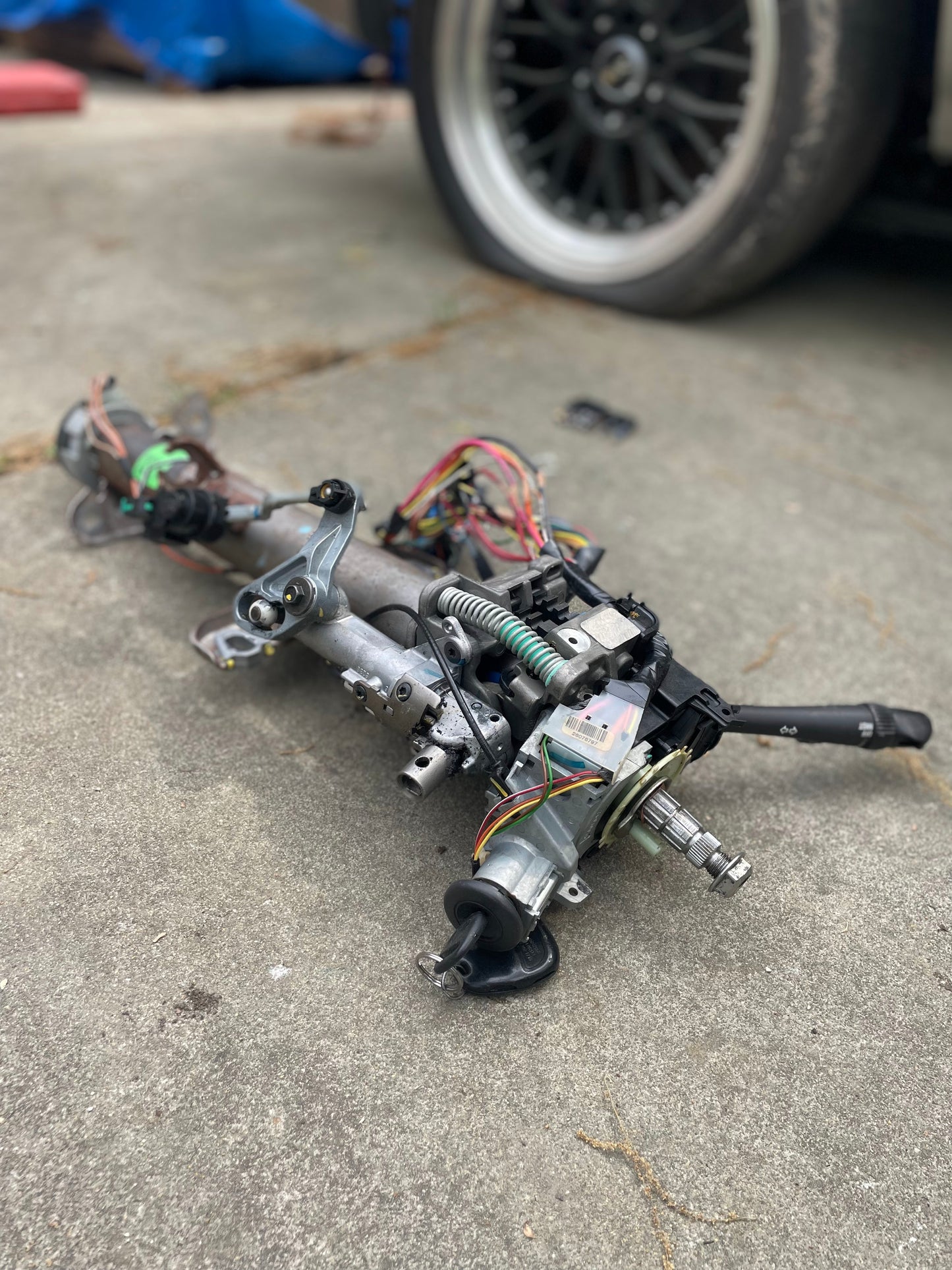 A General Motors OEM Steering Column w/KEY for 2000-2002 GM Full Size Trucks, from a model with automatic transmission, is shown with wiring lying on concrete near a parked Chevrolet Silverado, with its tire and rim visible in the background.