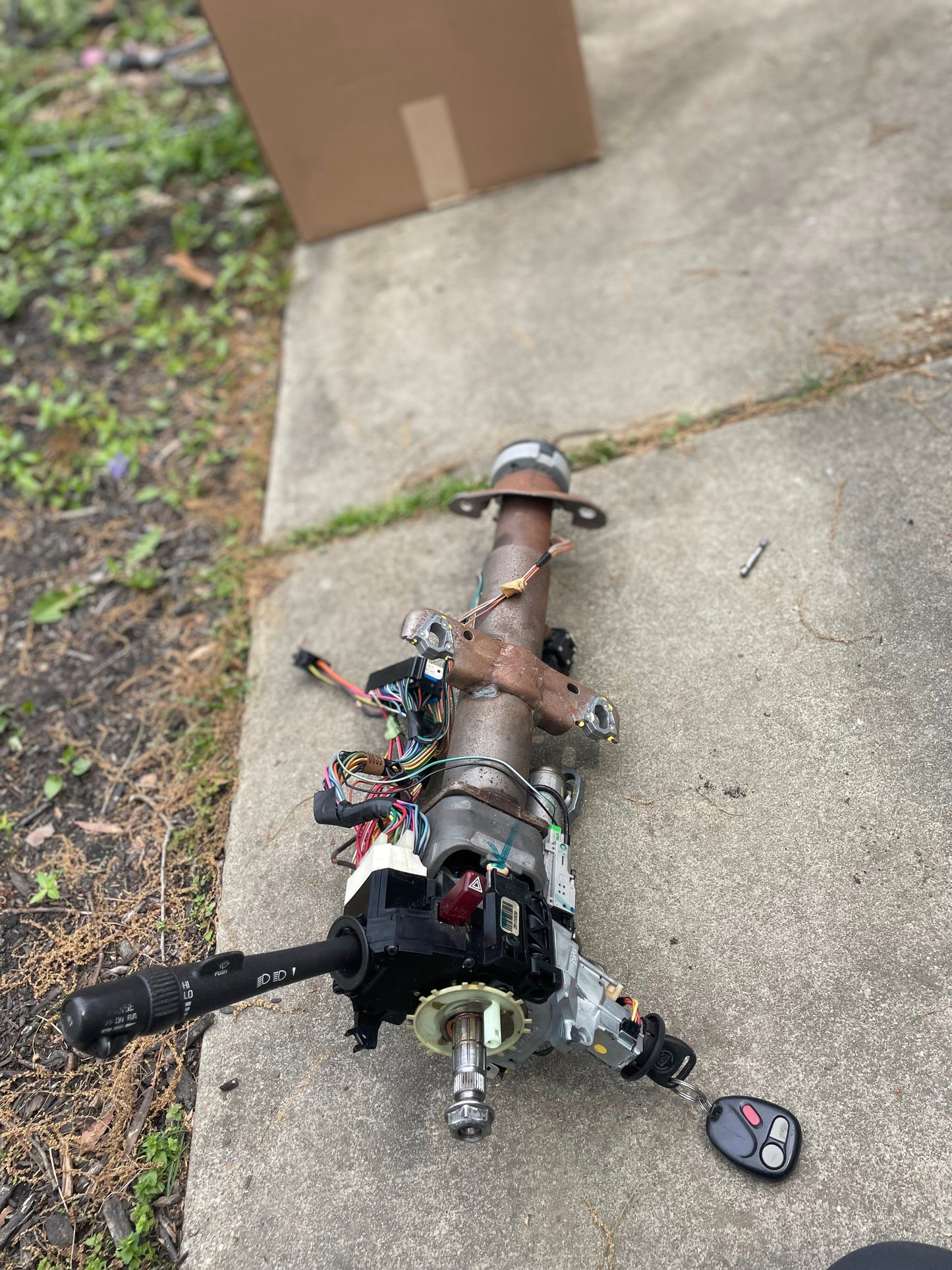 A General Motors OEM Steering Column with key for 2000-2002 GM Full Size Trucks rests outdoors on concrete, with wiring, controls, and key fob attached; grass and a cardboard box are visible in the background.