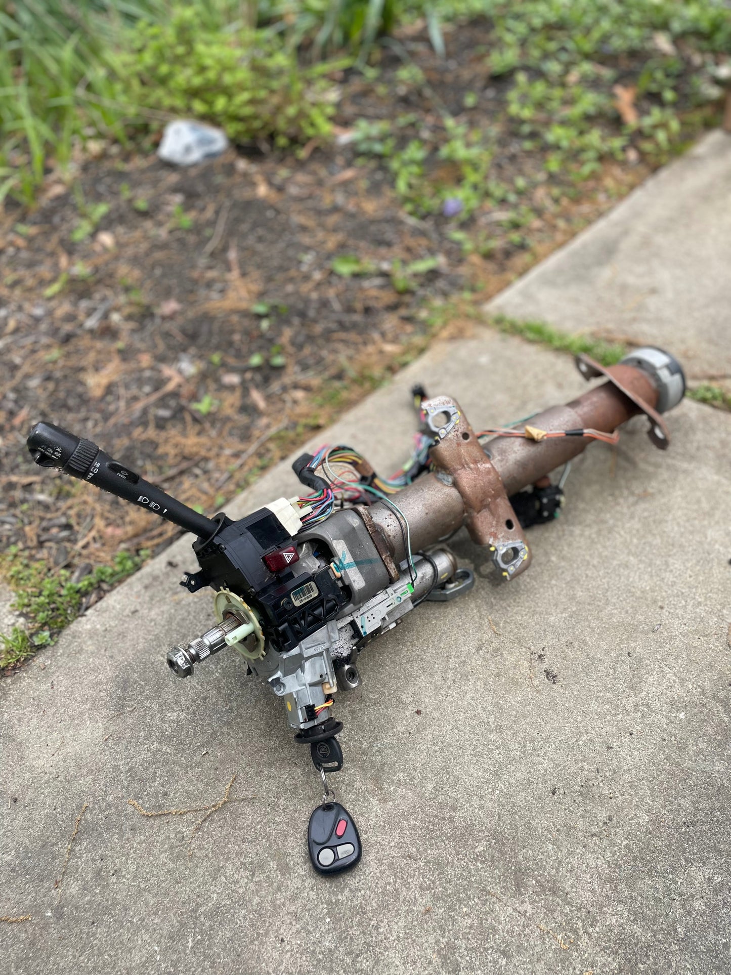 A disassembled General Motors OEM Steering Column w/KEY for 2000-2002 GM Full Size Trucks is shown with wiring and a remote key fob on a concrete surface outdoors, with grass and plants in the background.