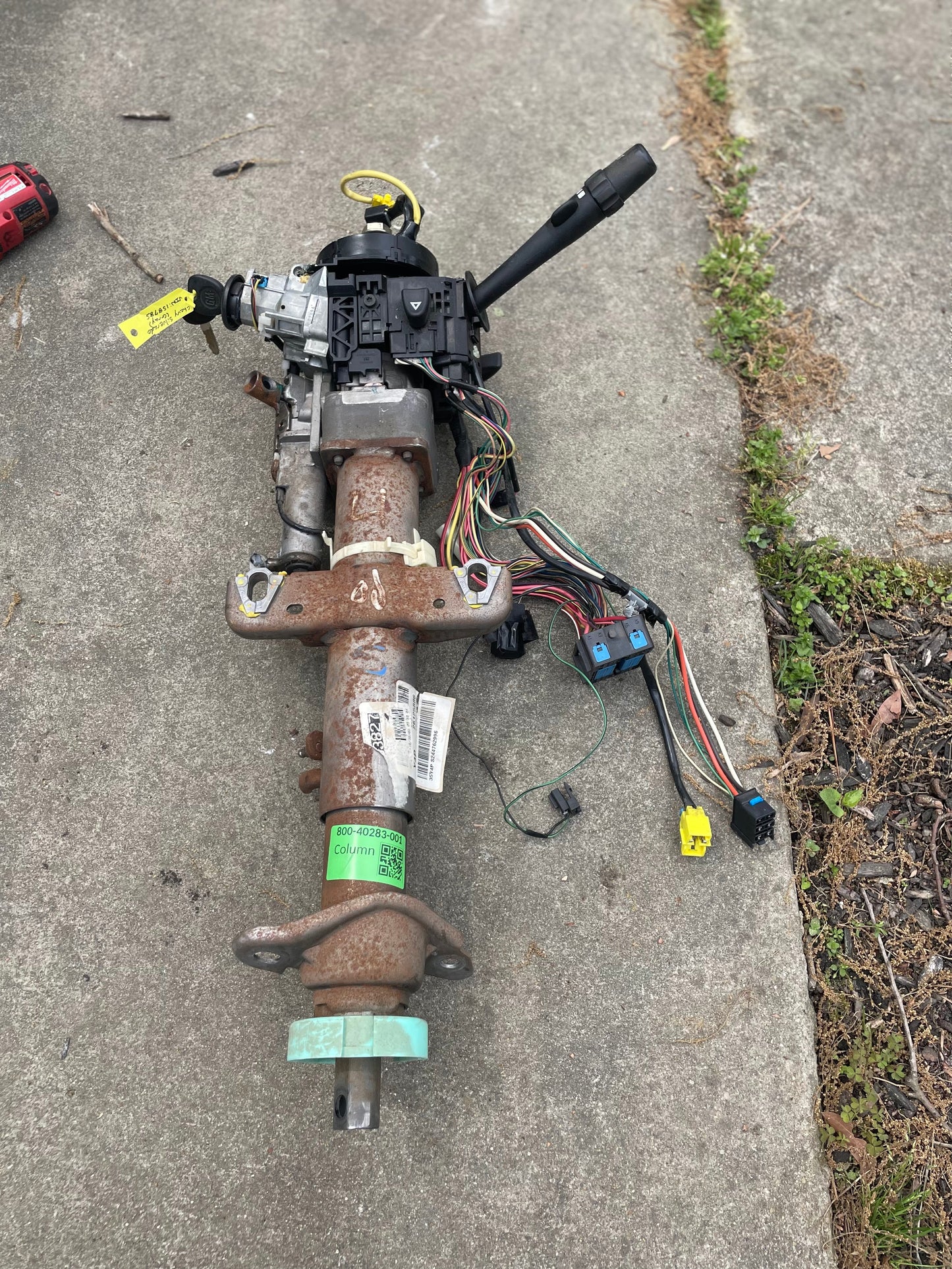 A General Motors OEM Steering Column w/KEY for 2003-2006 GM Full Size Trucks, including wiring, sensors, and control stalks for automatic transmission, is shown lying on a concrete surface next to some grass.