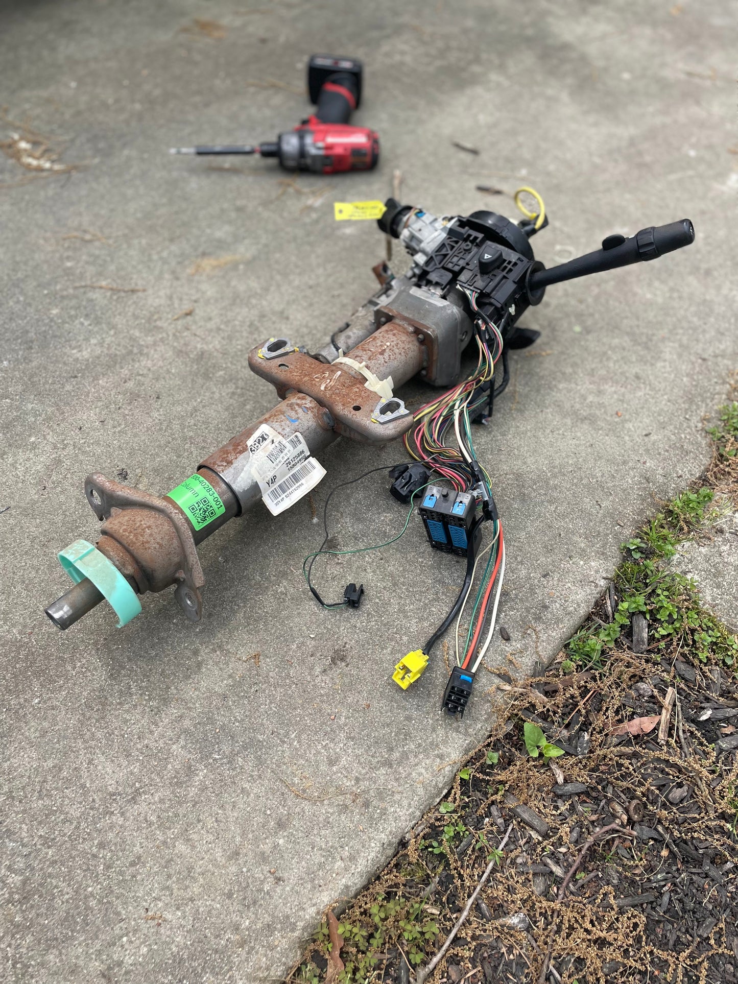 A General Motors OEM Steering Column w/KEY for 2003-2006 GM Full Size Trucks, complete with wiring and connectors, rests on a concrete surface. In the blurred background are a cordless drill, screwdriver, and some grass.