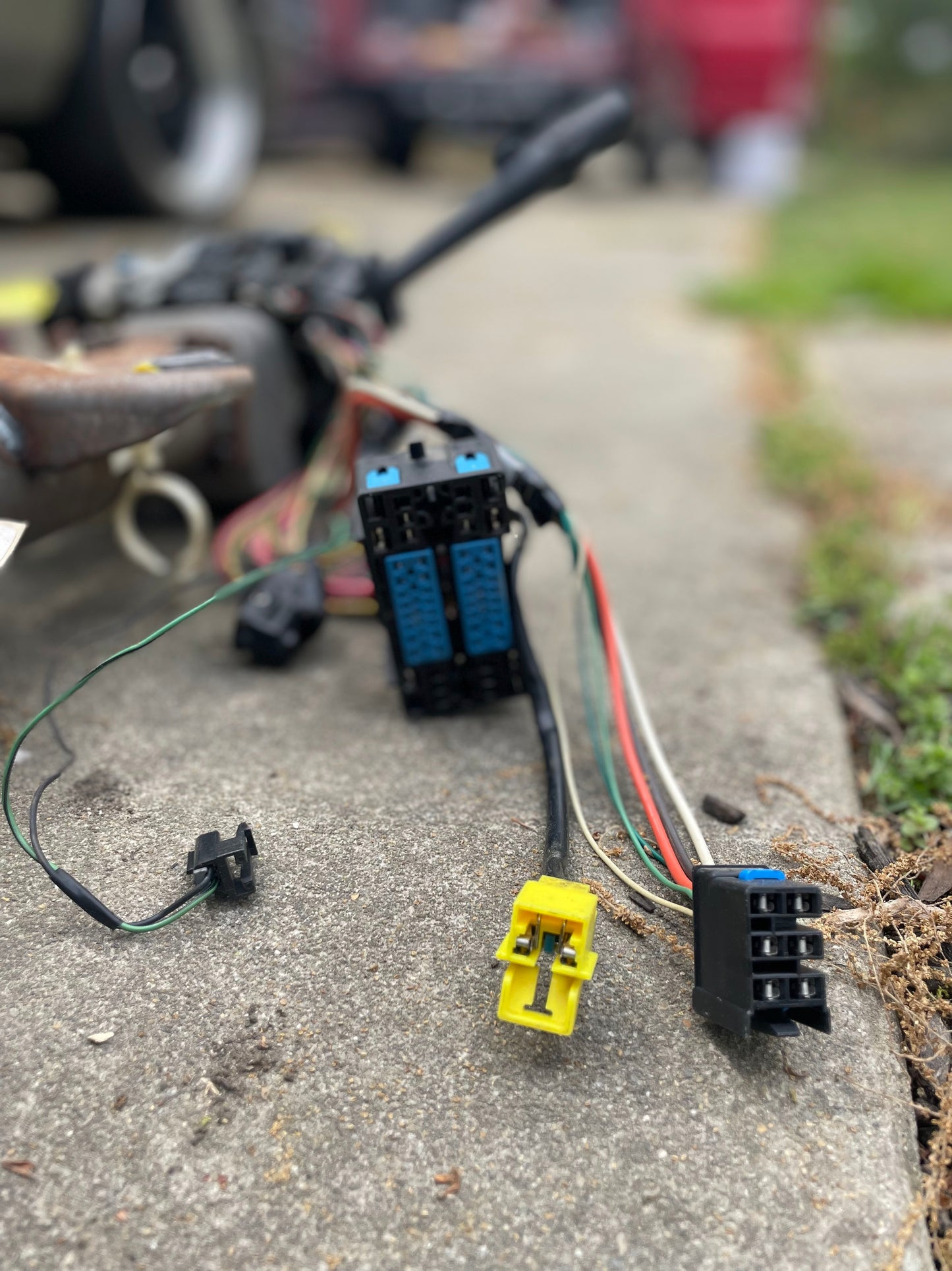 A close-up of colorful electrical connectors and wires on concrete outdoors, likely from a General Motors OEM Steering Column w/KEY for 2003-2006 GM Full Size Trucks, with blurred grass and vehicle parts in the background.