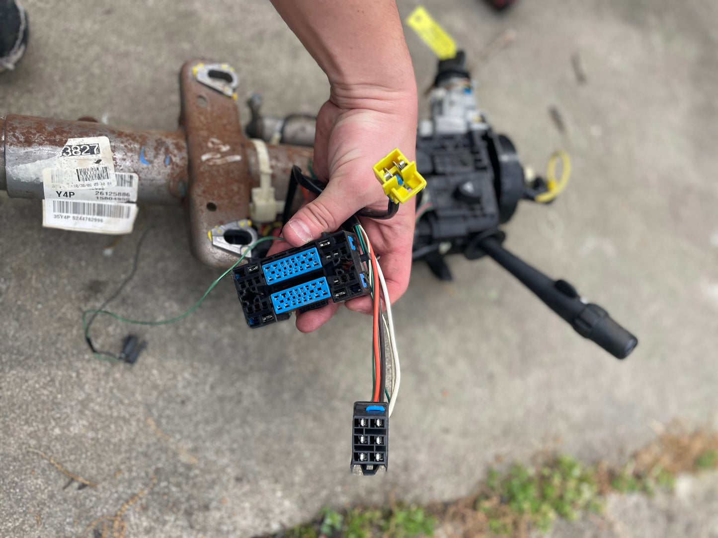 A hand holds several colorful electrical connectors attached to wires near a General Motors OEM Steering Column w/KEY for 2003-2006 GM Full Size Trucks, all resting on a concrete surface.