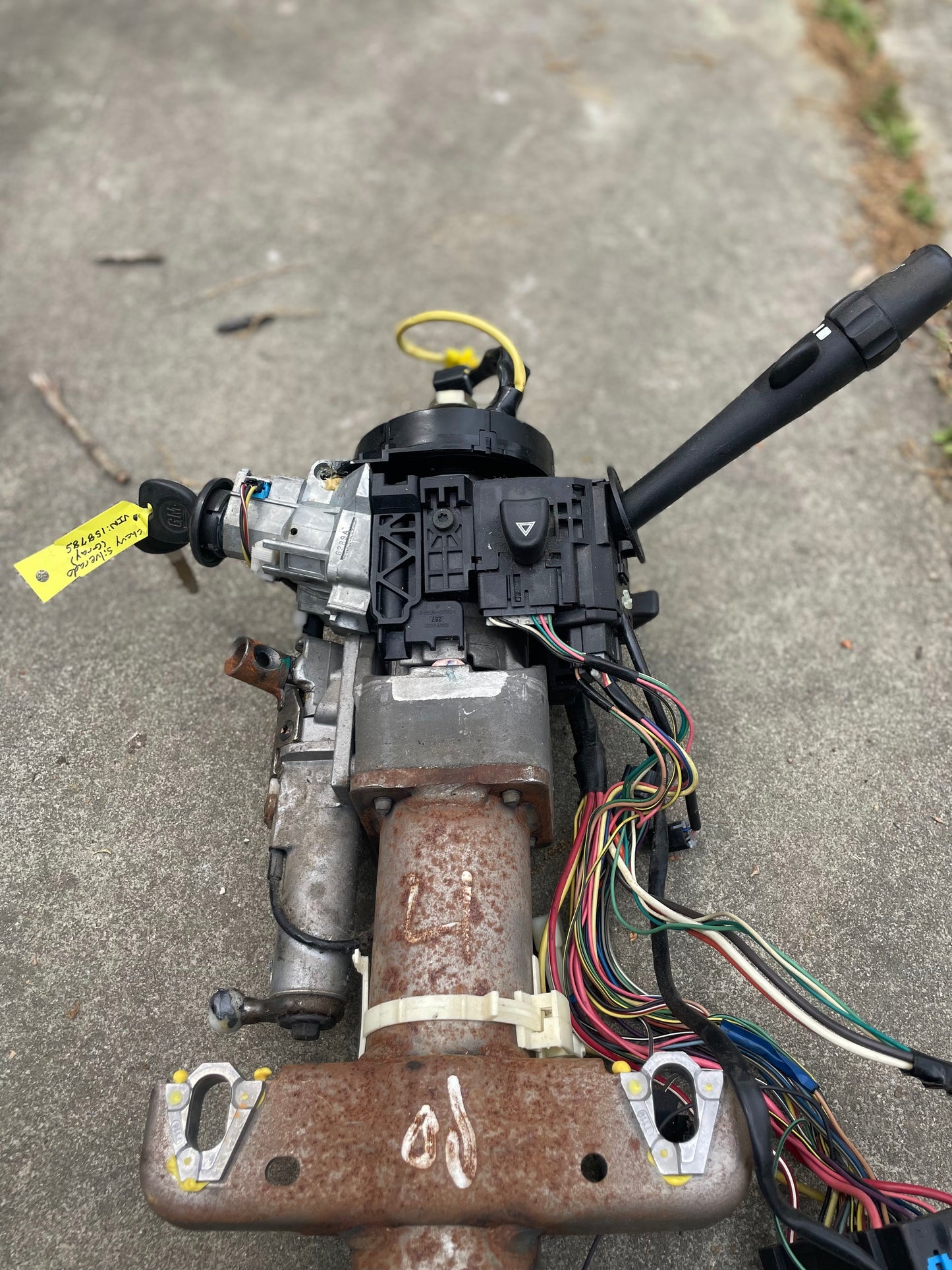 A close-up of a detached General Motors OEM Steering Column w/KEY for 2003-2006 GM Full Size Trucks, featuring exposed wires, rusted metal, turn signal lever, and automatic transmission indicator on concrete with grass at the corner.
