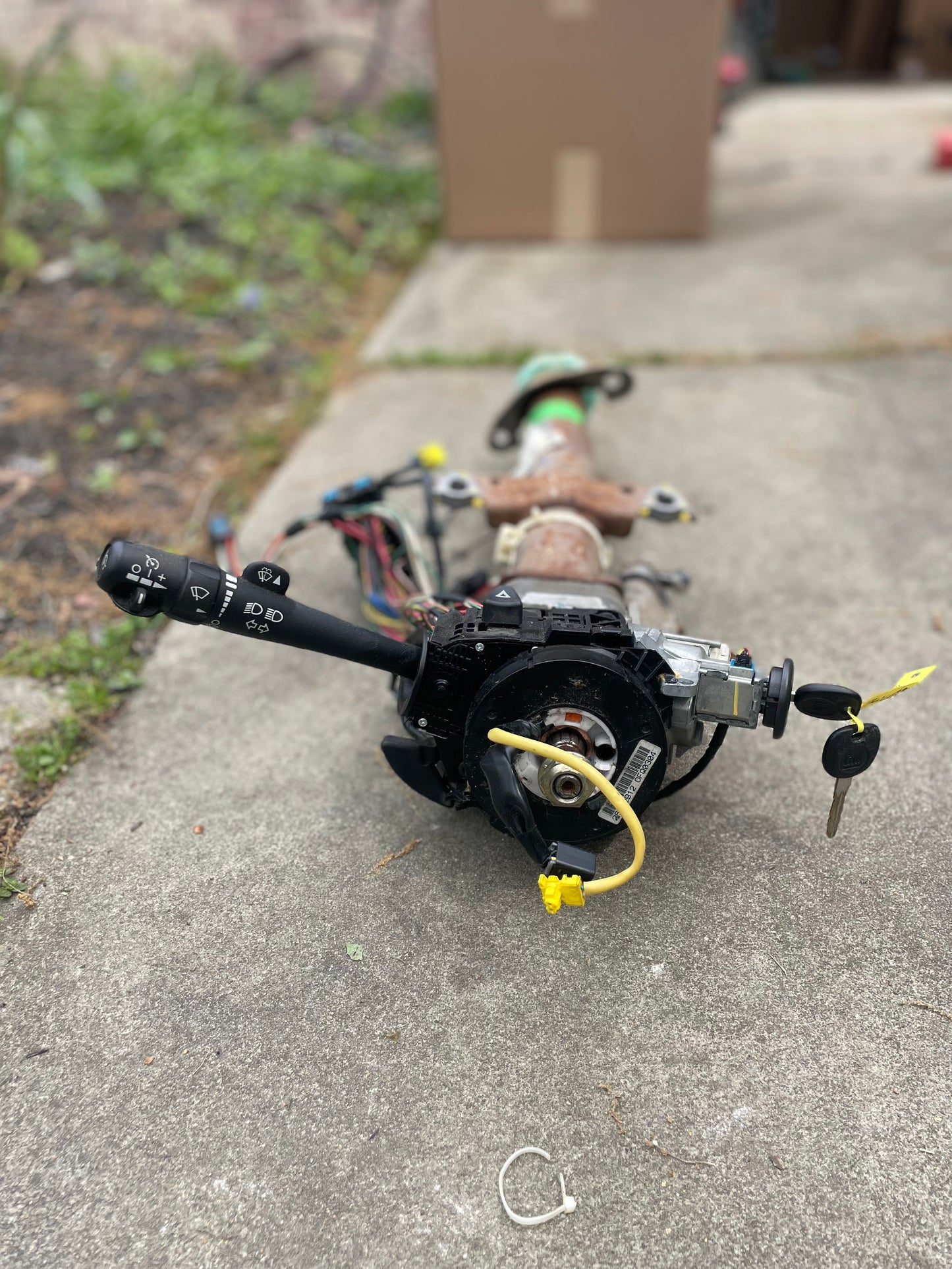 A General Motors OEM Steering Column w/KEY for 2003-2006 GM Full Size Trucks, featuring wiring and ignition switch, is shown on a concrete surface with grass and a cardboard box in the background.