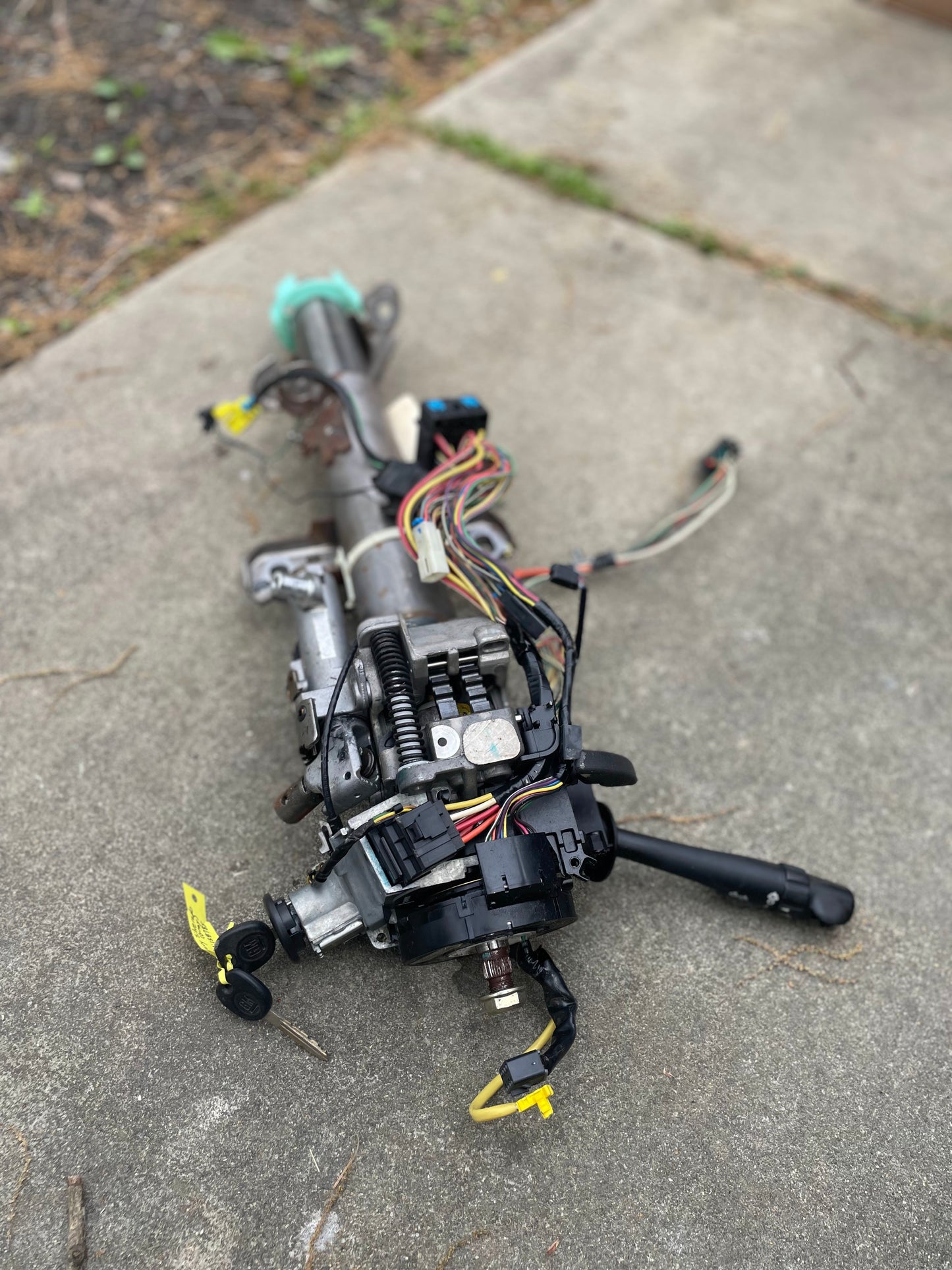 A General Motors OEM Steering Column w/KEY for 2003-2006 GM Full Size Trucks, featuring exposed wires, ignition, and levers for automatic transmission, is lying on a concrete surface outdoors.