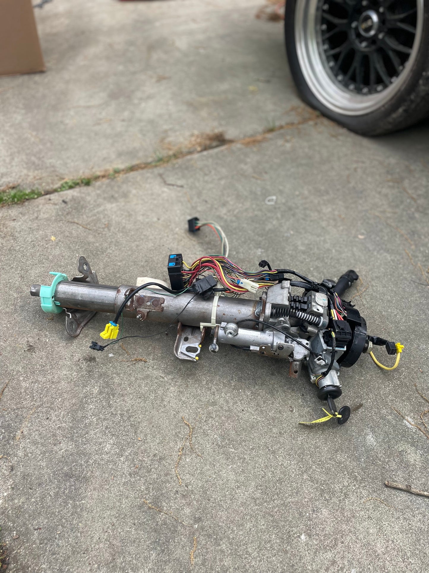 A General Motors OEM Steering Column w/KEY for 2003-2006 GM Full Size Trucks, with attached wiring and connectors, is placed on a concrete surface beside a black Chevrolet Silverado wheel.
