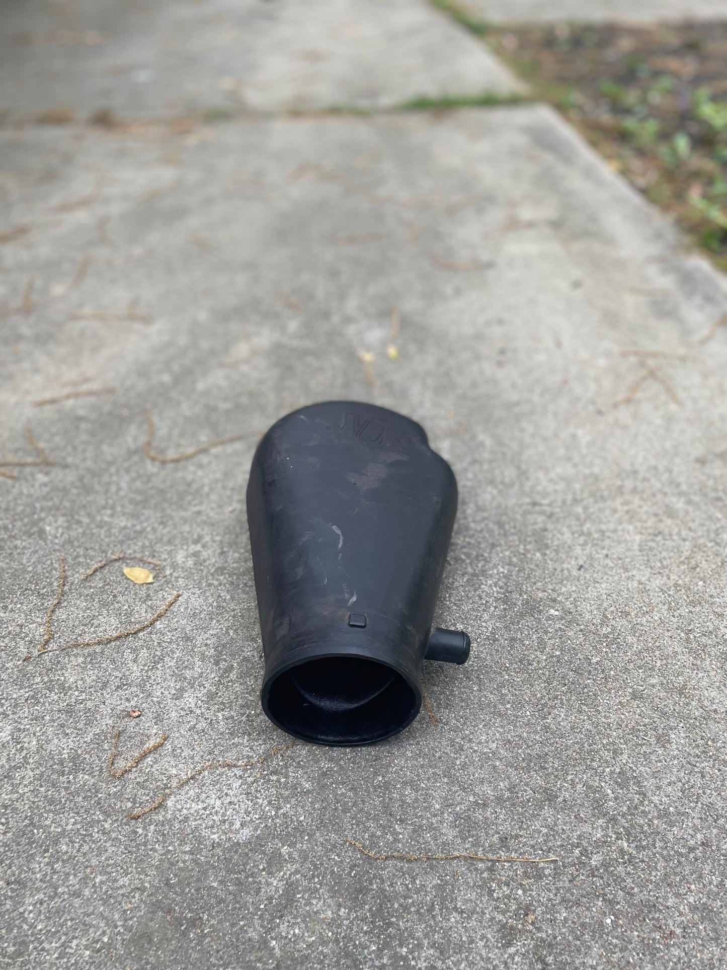 A black plastic General Motors OEM Cold Air Intake Hat 25147235 for a 4.3L 1996-2005 Chevrolet S10, Blazer, and more is on a concrete sidewalk with dry twigs and greenery in the background.
