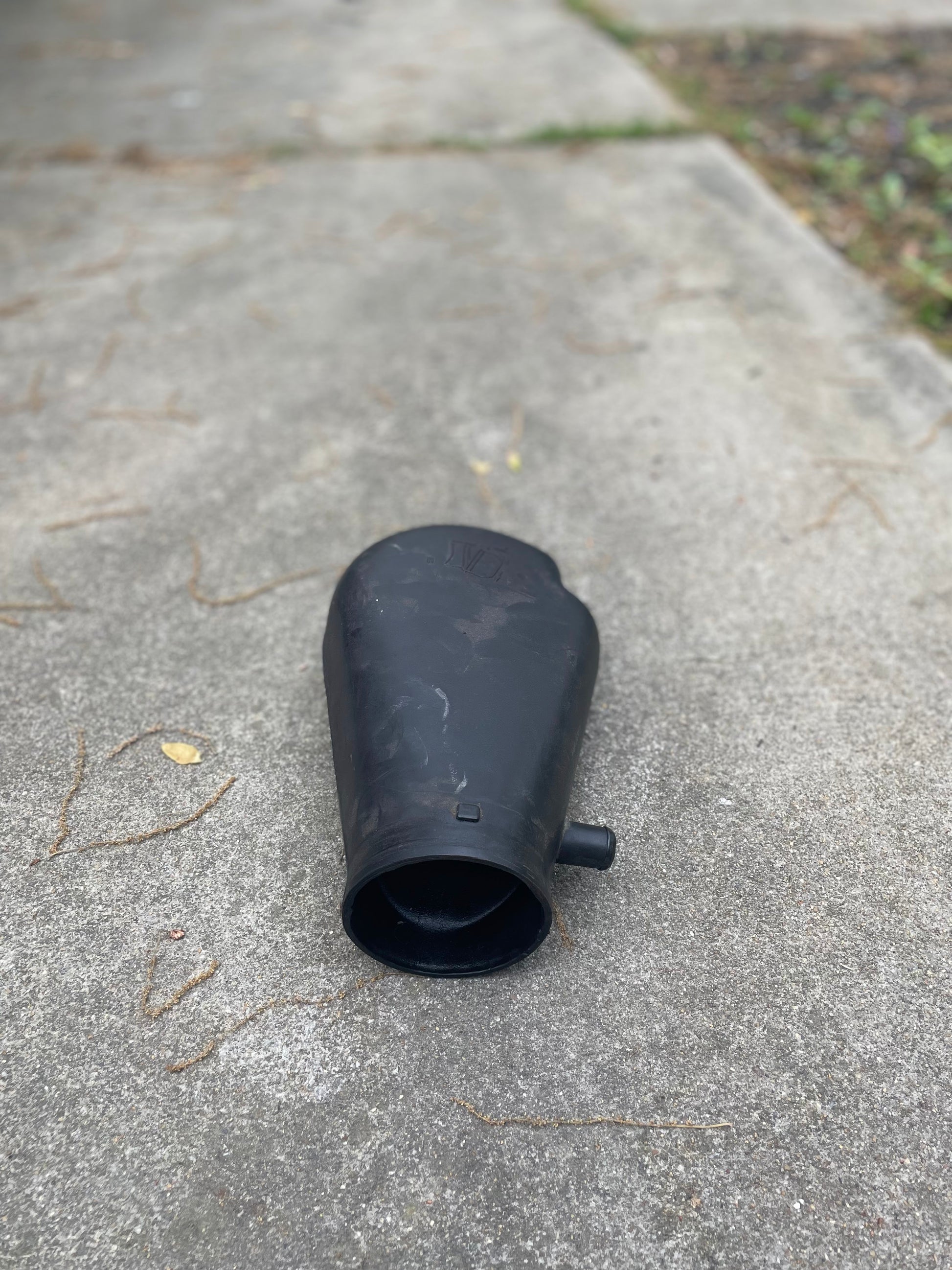 A black plastic General Motors OEM Cold Air Intake Hat 25147235 for a 4.3L 1996-2005 Chevrolet S10, Blazer, and more is on a concrete sidewalk with dry twigs and greenery in the background.