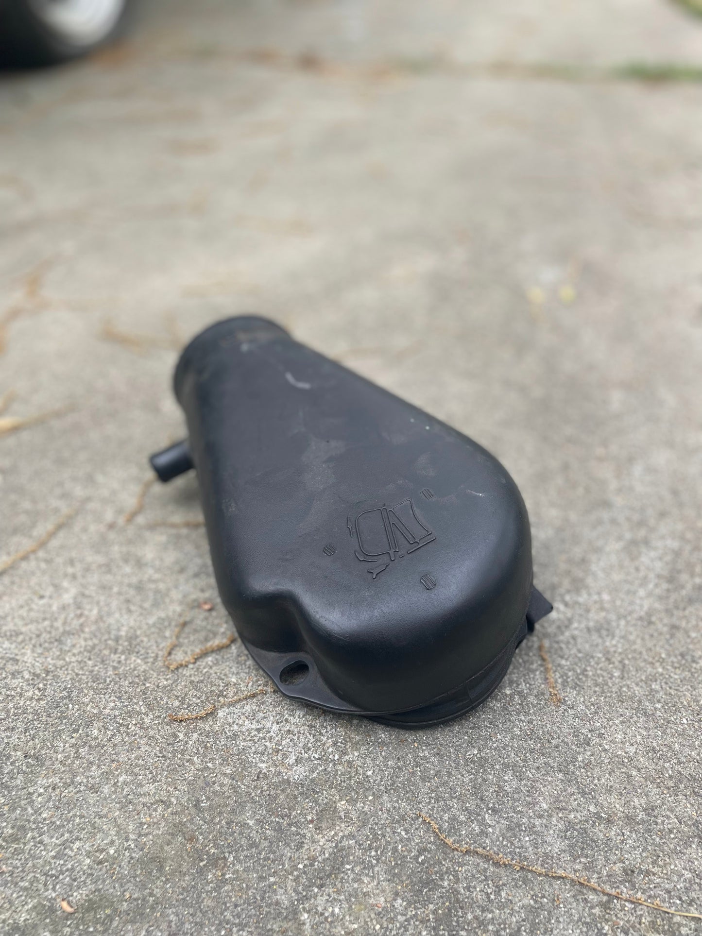 A black General Motors OEM Cold Air Intake Hat (part 25147235) for 4.3L 1996–2005 Chevrolet S10, Blazer, and more sits on concrete with dried grass nearby. The background is slightly blurred.