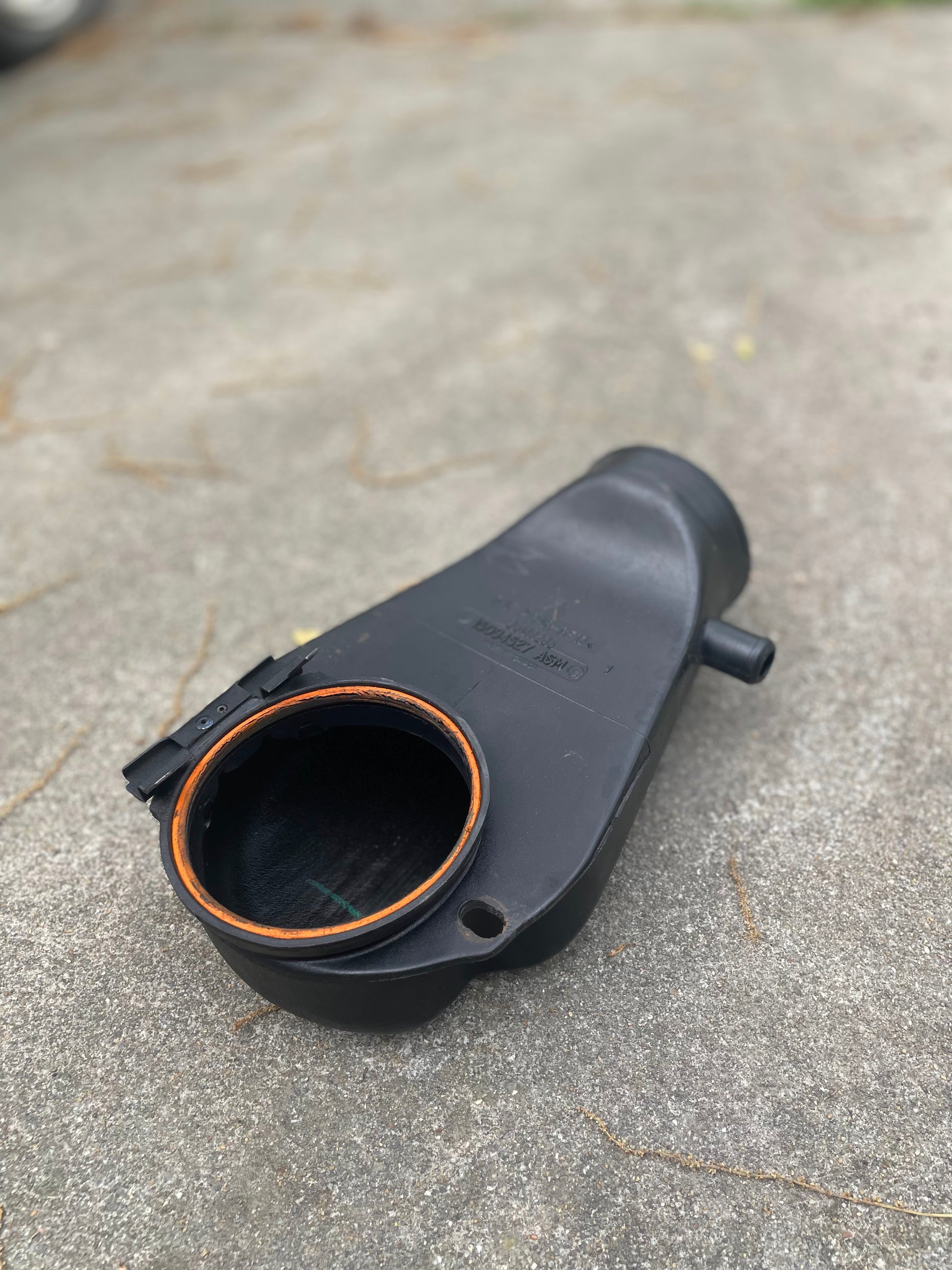A black General Motors OEM Cold Air Intake Hat 25147235 with an orange gasket for a 4.3L 1996-2005 Chevrolet S10, Blazer, and more lies on a textured concrete surface outdoors, open end facing the camera.