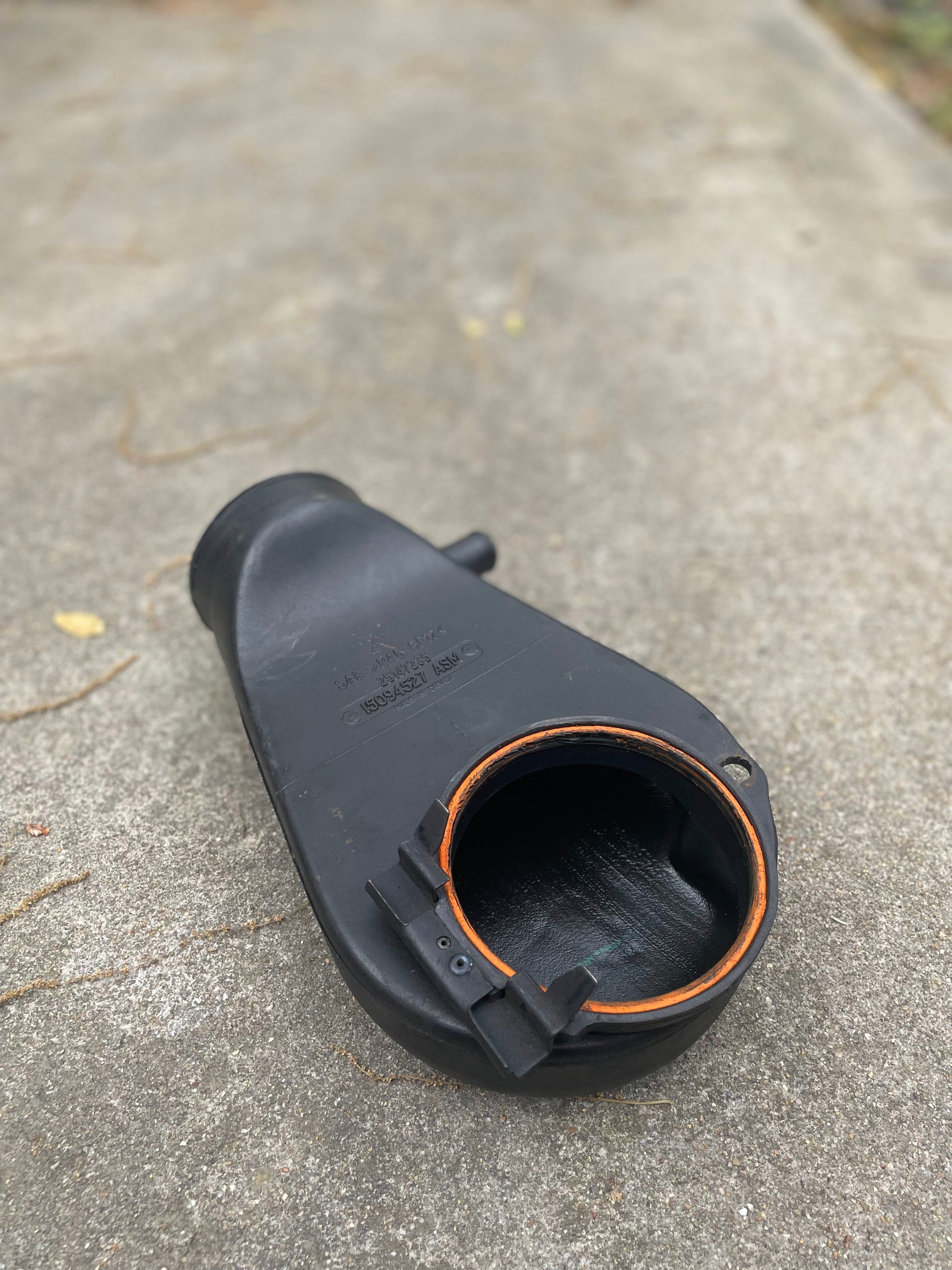 A black General Motors OEM Cold Air Intake Hat 25147235 with an orange seal sits on concrete, its open end in focus. Designed for a 4.3L 1996–2005 Chevrolet S10, Blazer, and more; perfect for throttle body adapter upgrades.