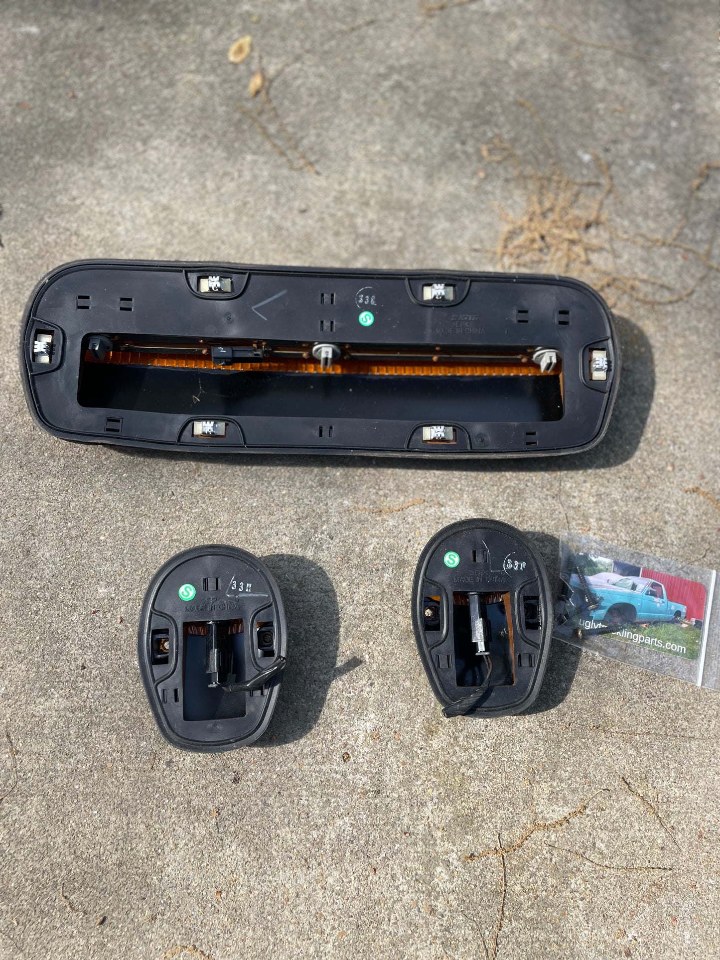 Three black plastic General Motors OEM roof clearance light lamp lenses (left, right, center) for 2000–2007 Chevrolet Silverado, Suburban, and more are arranged on concrete. Two are labeled L and R. A business card shows a green truck.