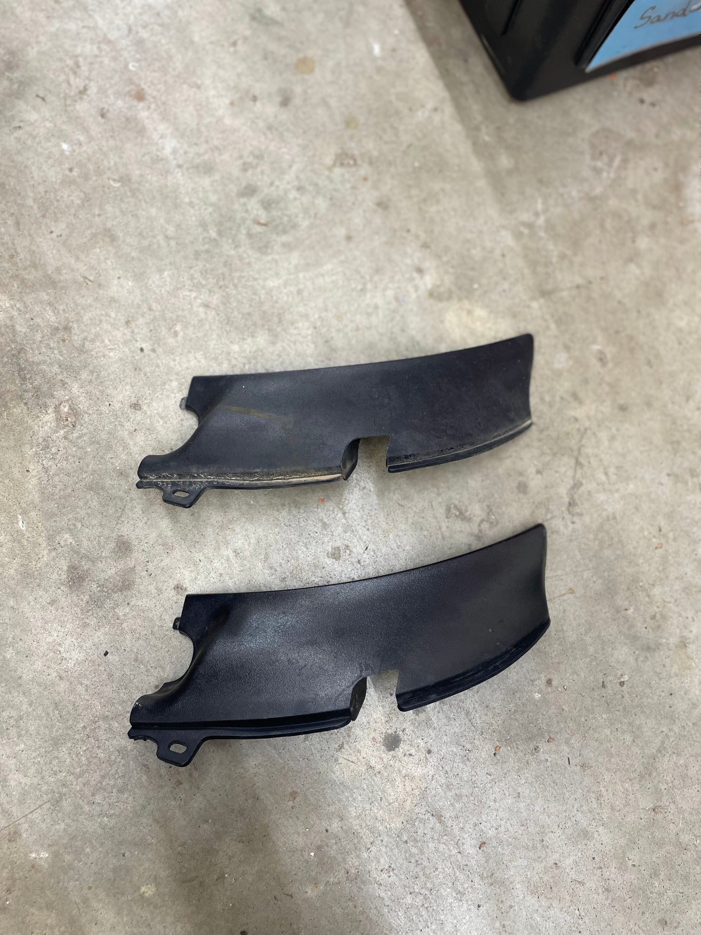 Two black OEM General Motors wiper cowl covers, left driver trim pieces with texture for 1994-2005 Chevrolet S10, Blazer, and more, lie parallel on a concrete floor. They appear slightly worn; a black container is partly visible in the top right corner.
