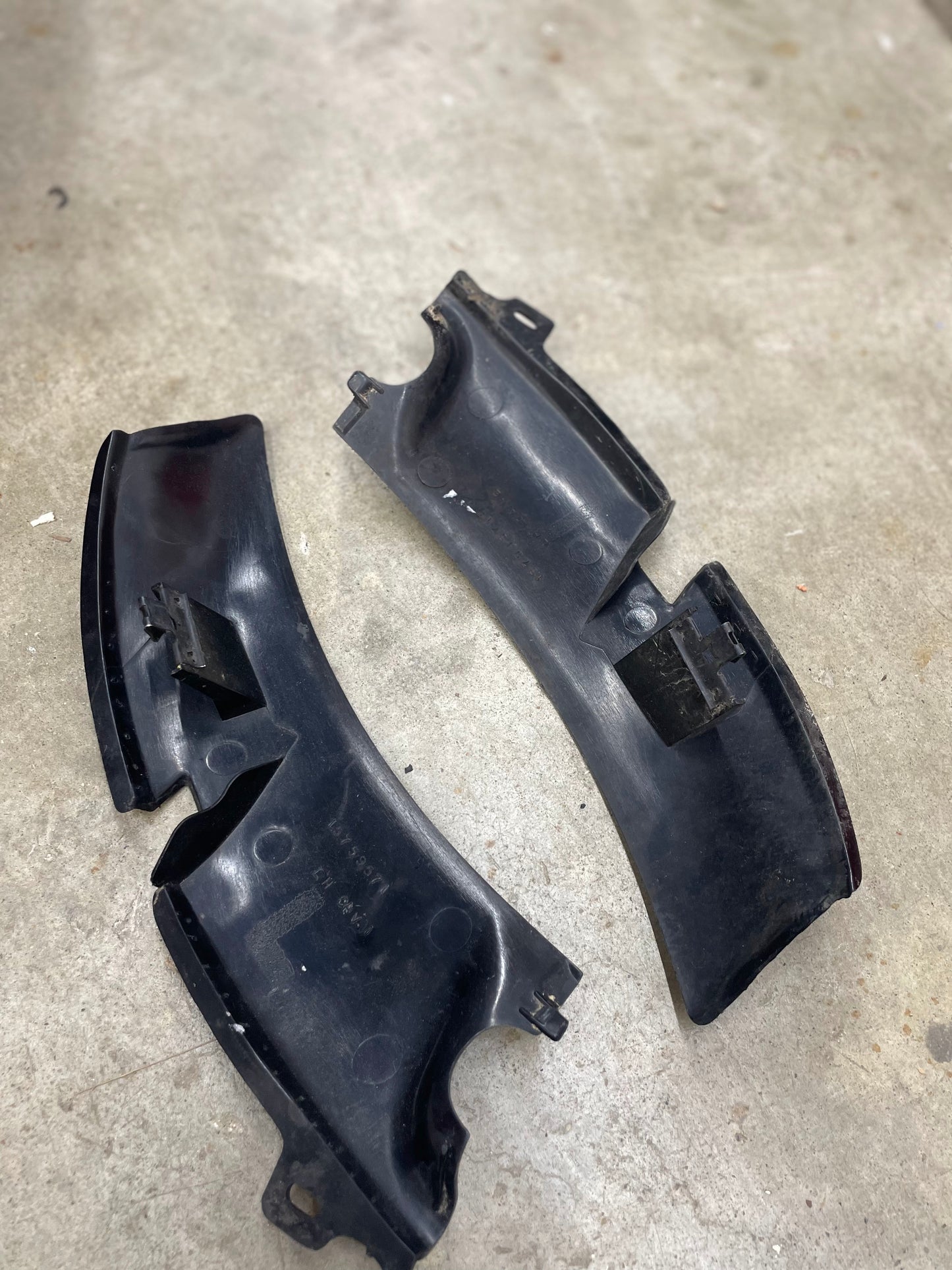 A pair of OEM Wiper Cowl Cover Left Driver Trims with texture by General Motors for 1994-2005 Chevrolet S10, Blazer, and more, are lying on a concrete floor. Slightly worn, they feature attachment clips and ridges.