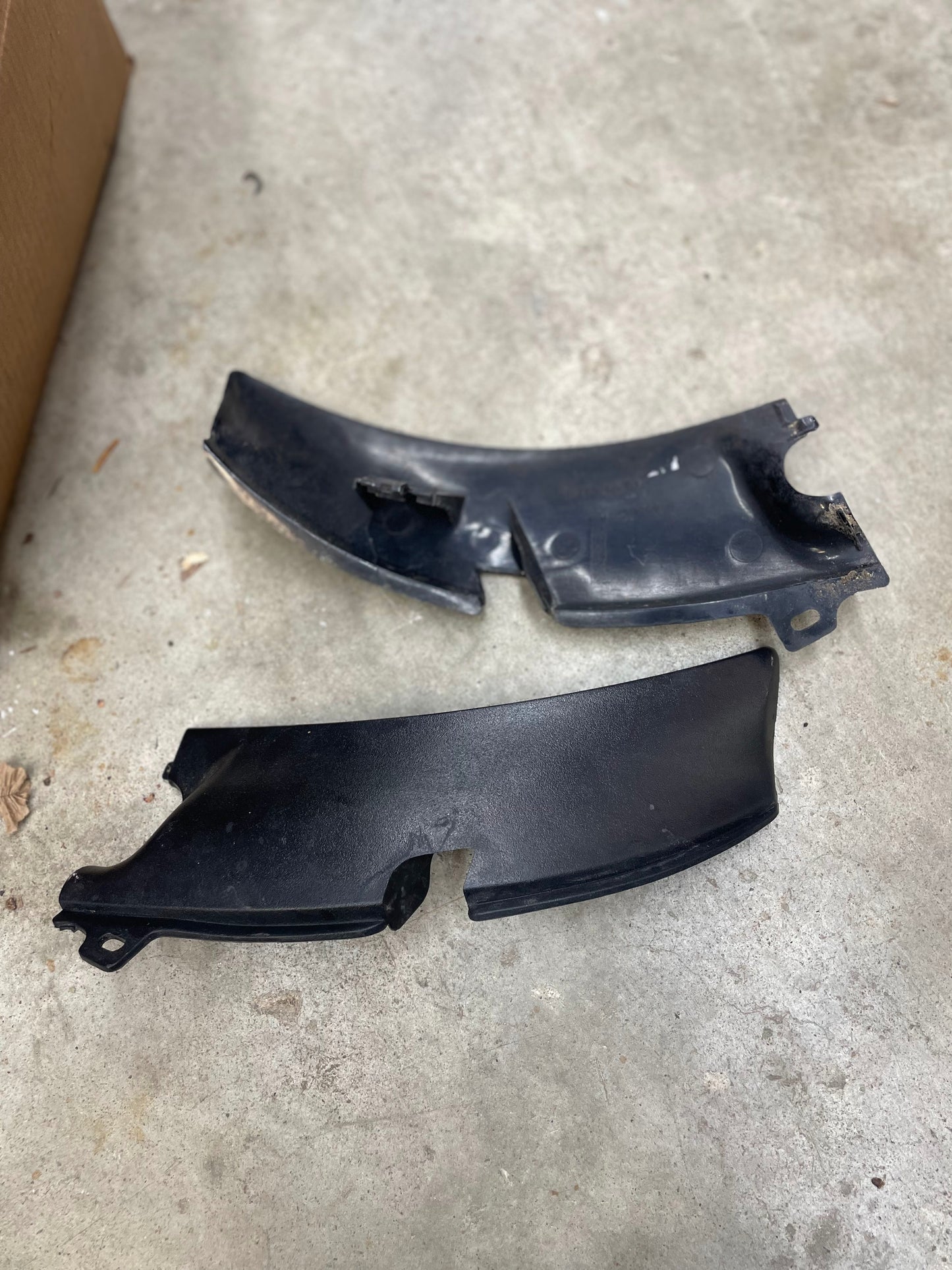 Two black plastic car parts, including a cracked General Motors OEM Wiper Cowl Cover Left Driver Trim with texture for 1994-2005 Chevrolet S10 and Blazer, are lying on a concrete floor beside a cardboard box.