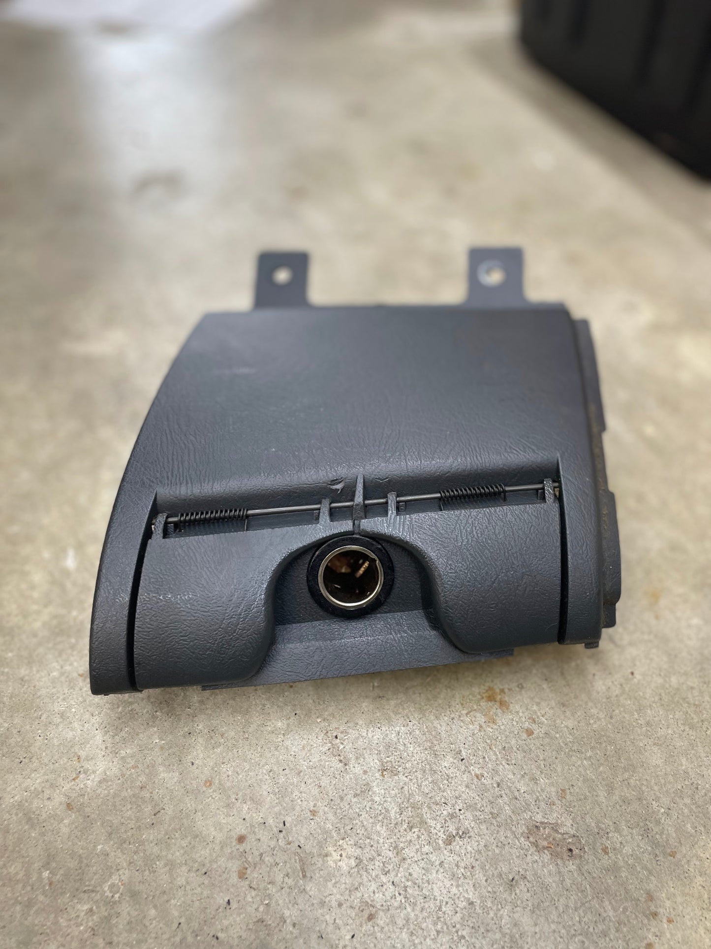A dark gray OEM 3 Port 12V Aux Panel Ash Tray Delete by General Motors, for 1998-2000 GMC Envoy, suited for charcoal interiors. Features lock cylinder and two top mounting brackets; shown on a concrete floor.