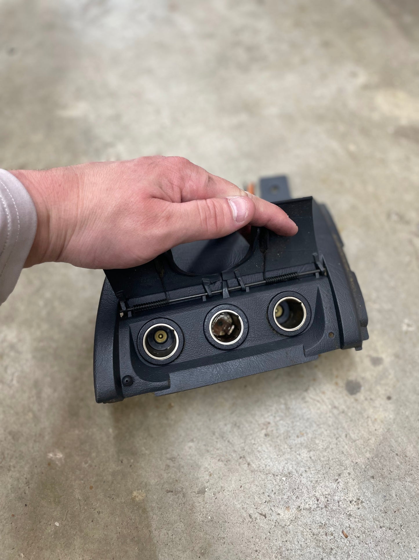 A hand lifts the cover of a General Motors OEM 3 Port 12V Aux Panel Ash Tray Delete in dark gray, designed for 1998–2000 GMC Envoy, revealing three circular ports set within charcoal interior trim.